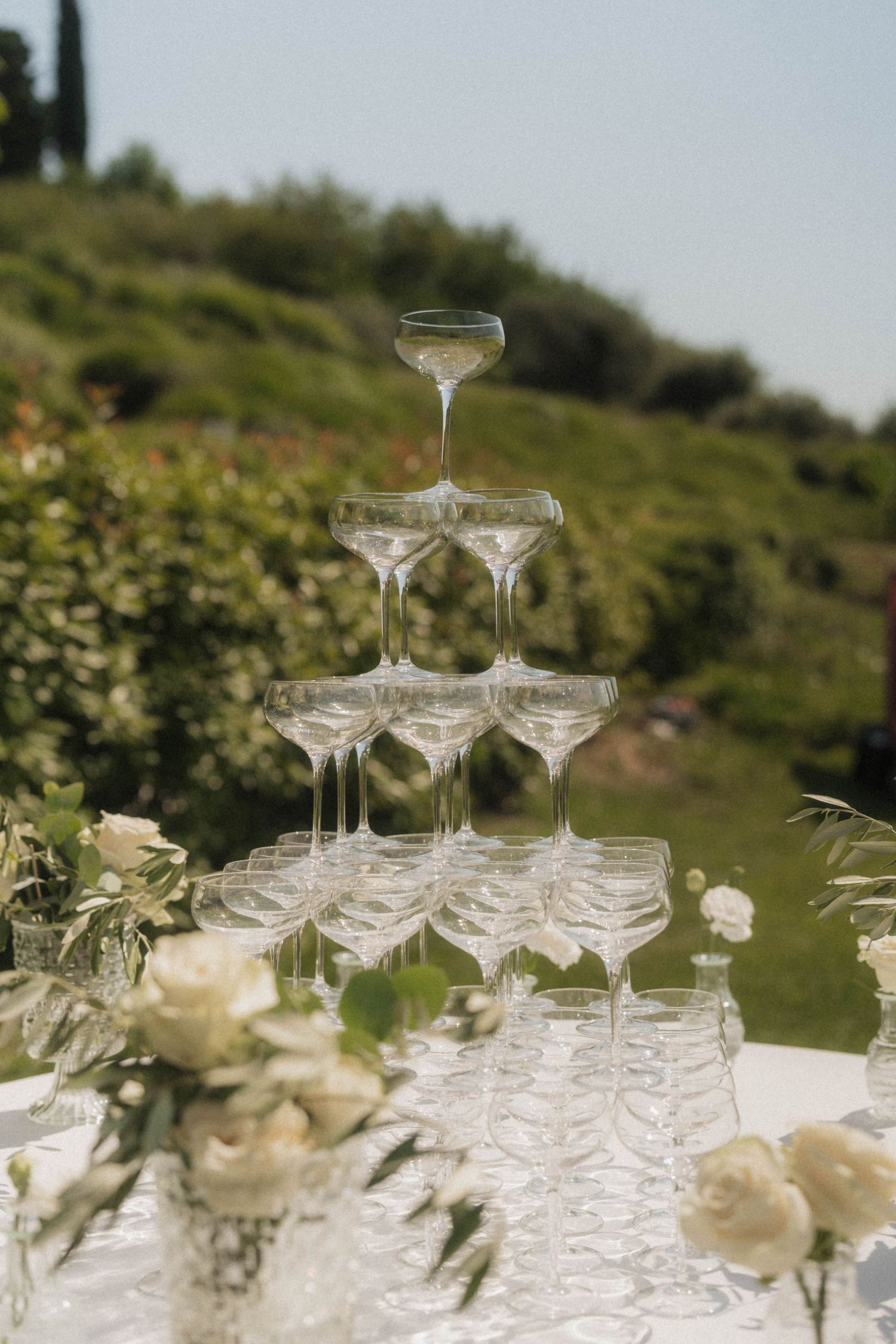 Pyramid of champagne coupe glasses on a white table with white roses.