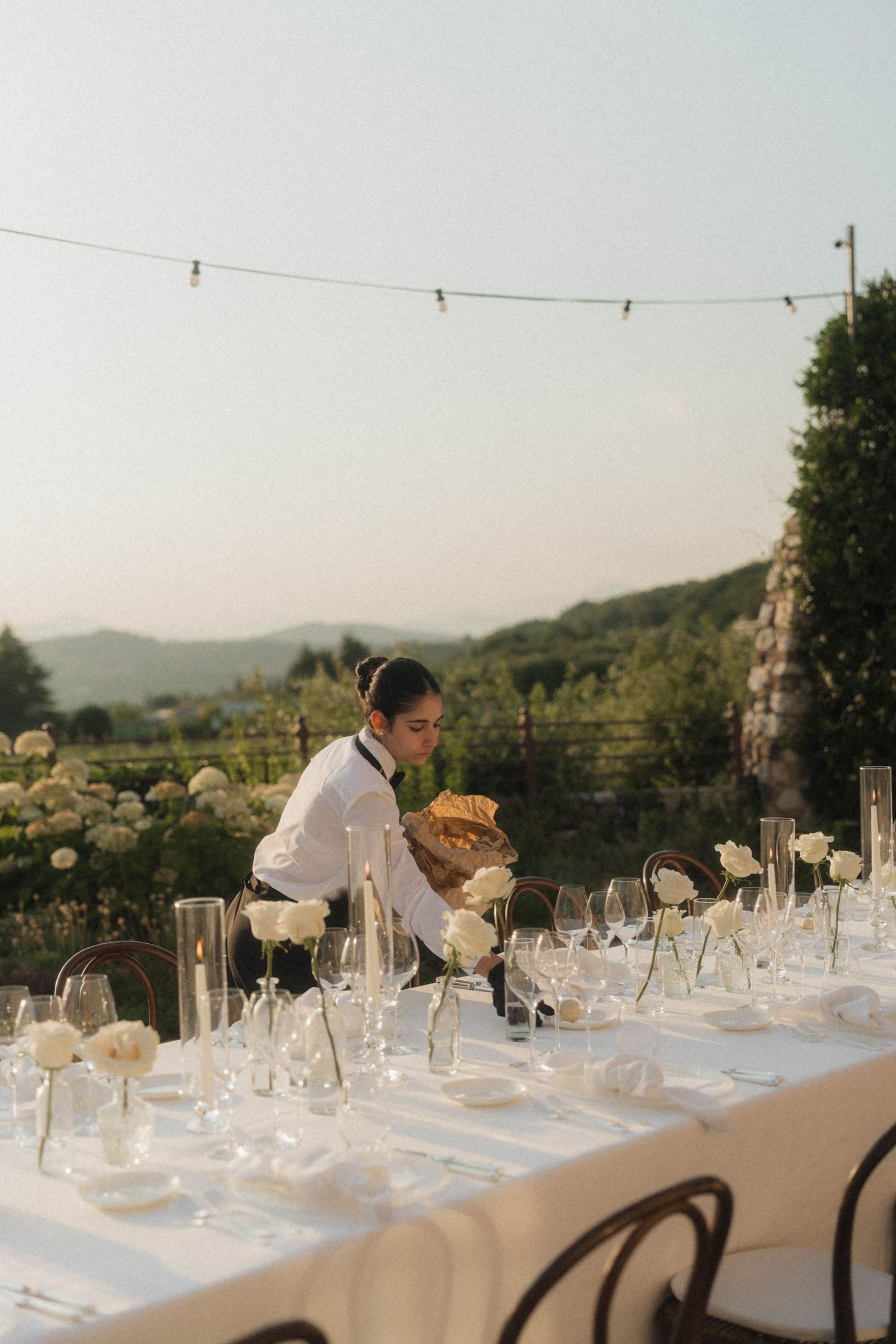 Waiter setting a long elegant outdoor wedding reception table at dusk.