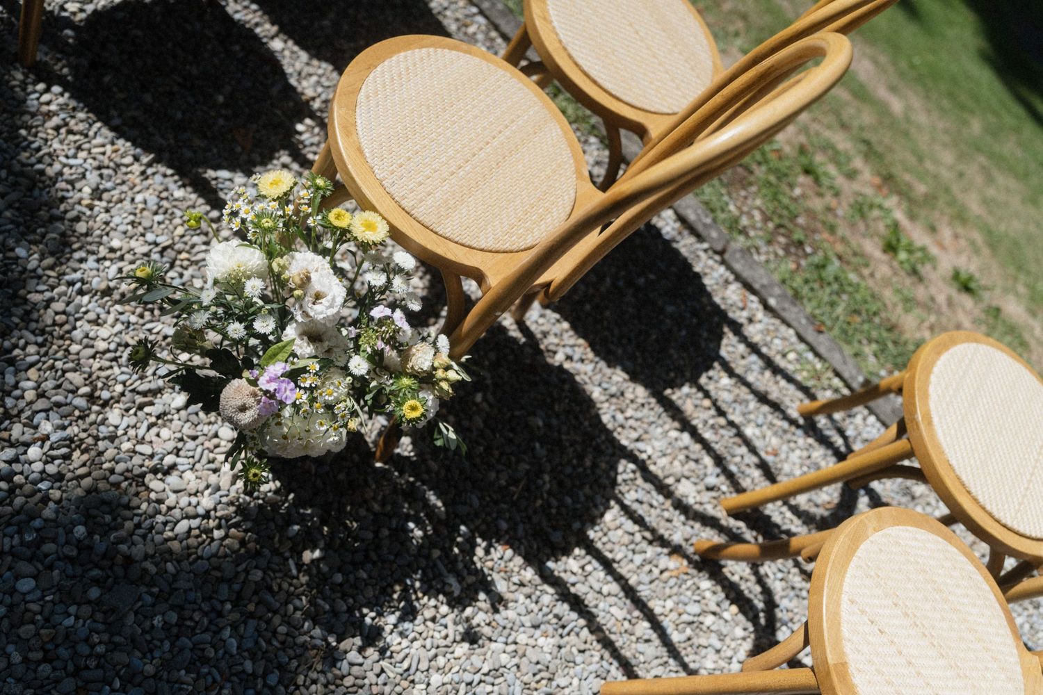 Elegant rattan chairs arranged outdoors on gravel with a delicate wildflower bouquet for a garden wedding ceremony.