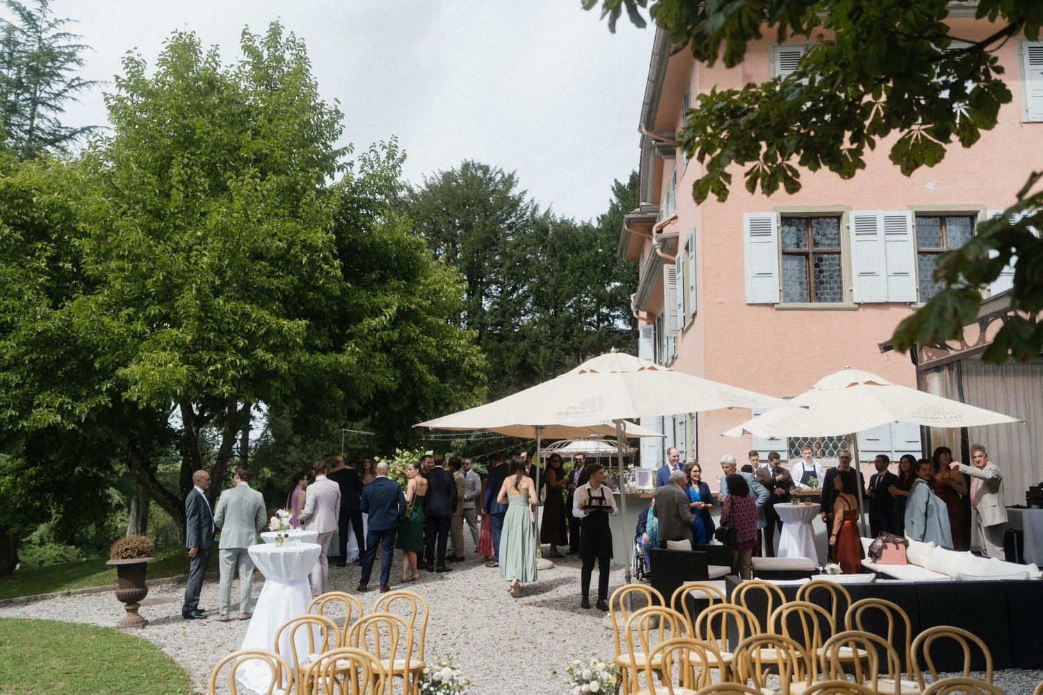 Outdoor wedding reception at a villa with guests mingling under white umbrellas surrounded by lush trees.