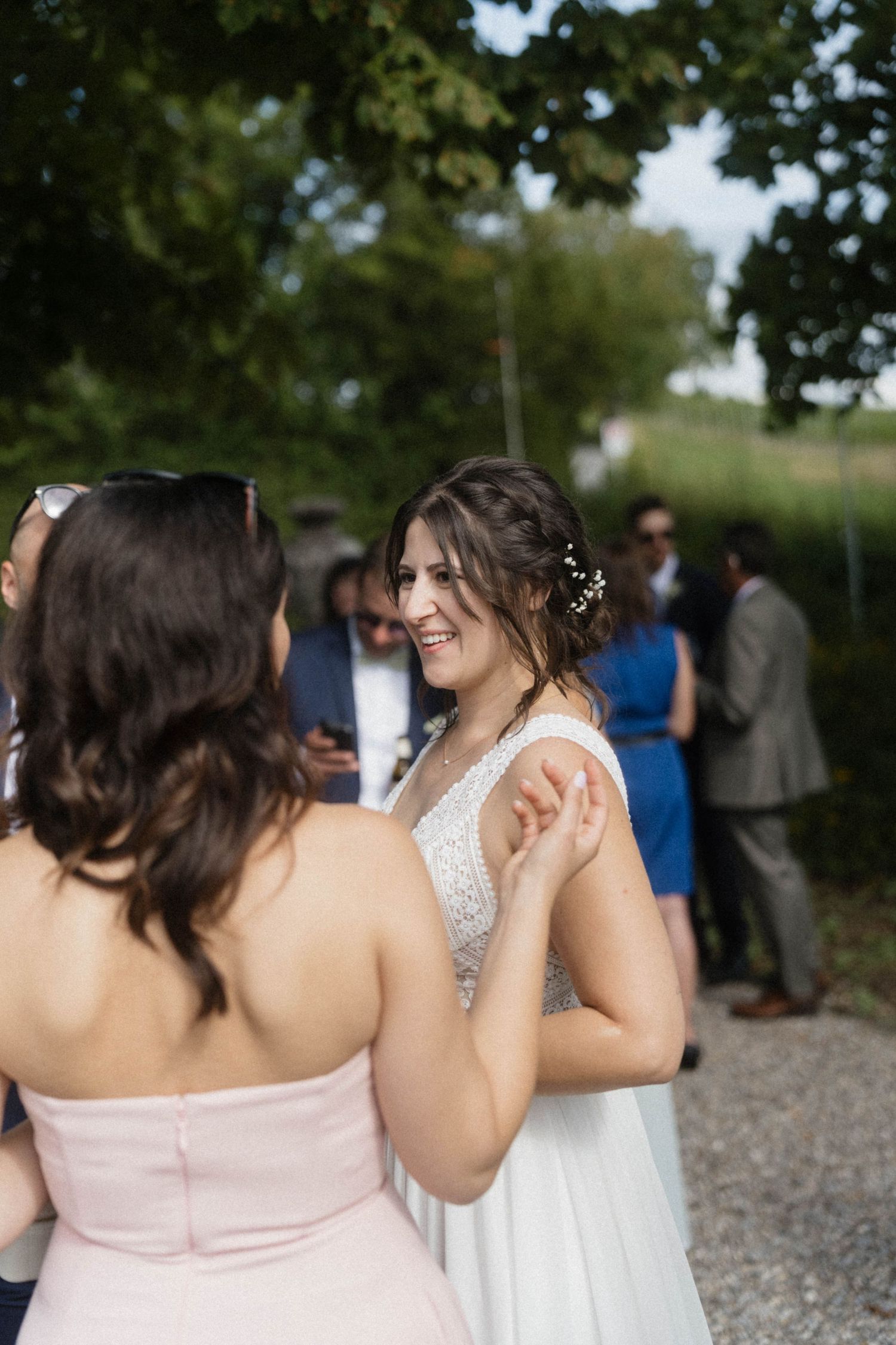 Joyful bride in a white beaded gown laughing with a guest in a pink dress at an outdoor wedding.