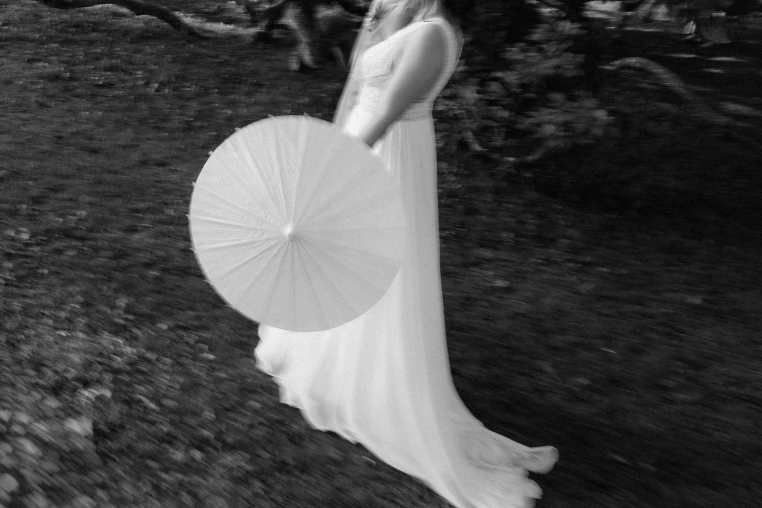 Black and white photo of bride in flowing white gown holding a parasol umbrella outdoors.