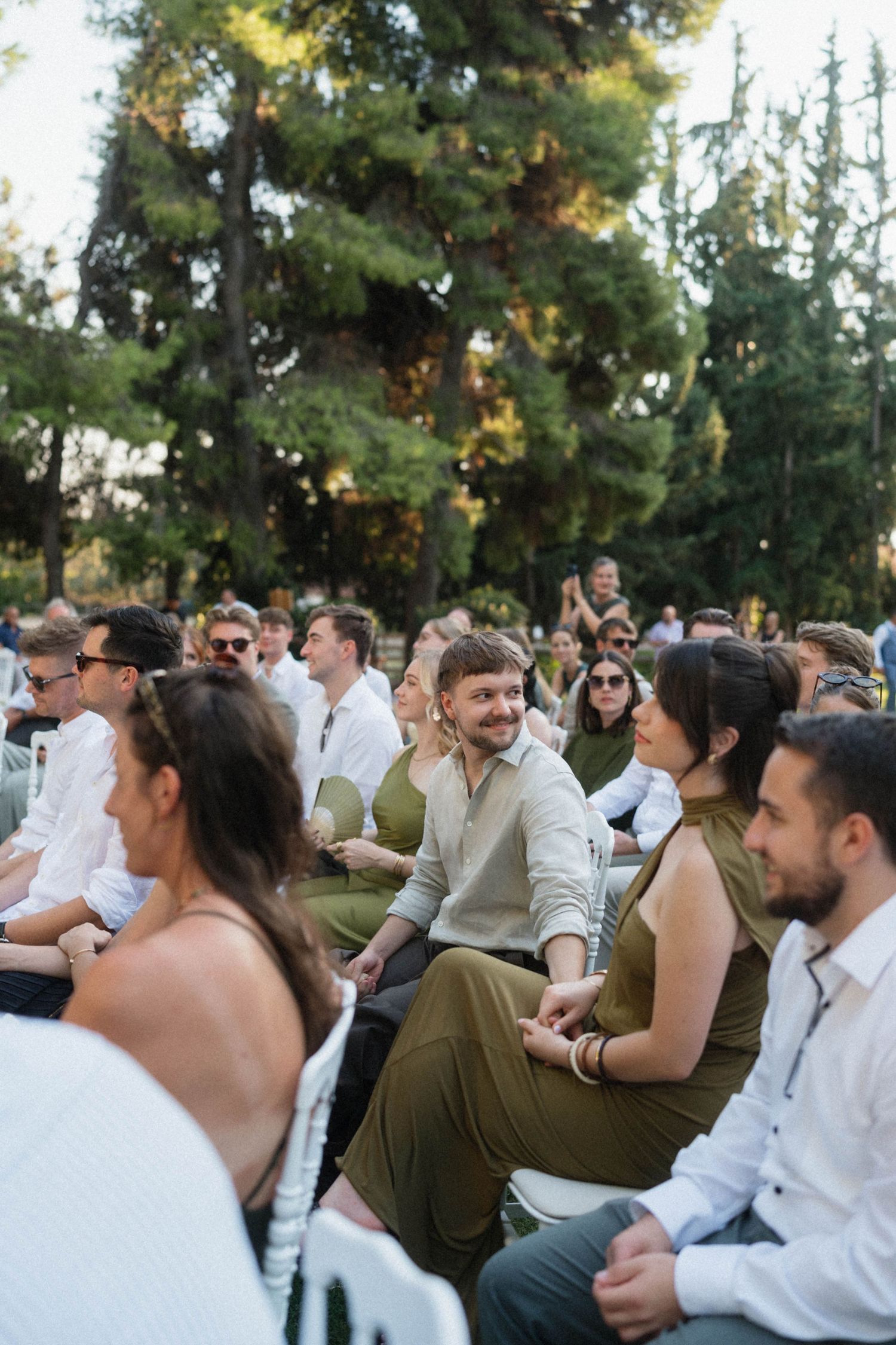 Wedding guests seated in rows of white chairs smile during an outdoor ceremony surrounded by lush green trees.