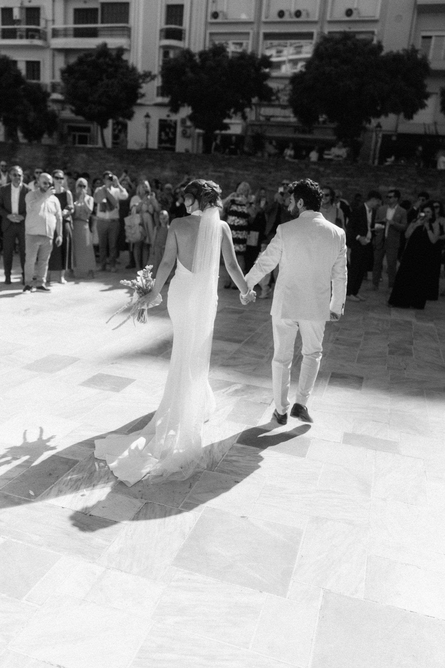 Black and white photo of bride and groom holding hands walking toward wedding guests outdoors.