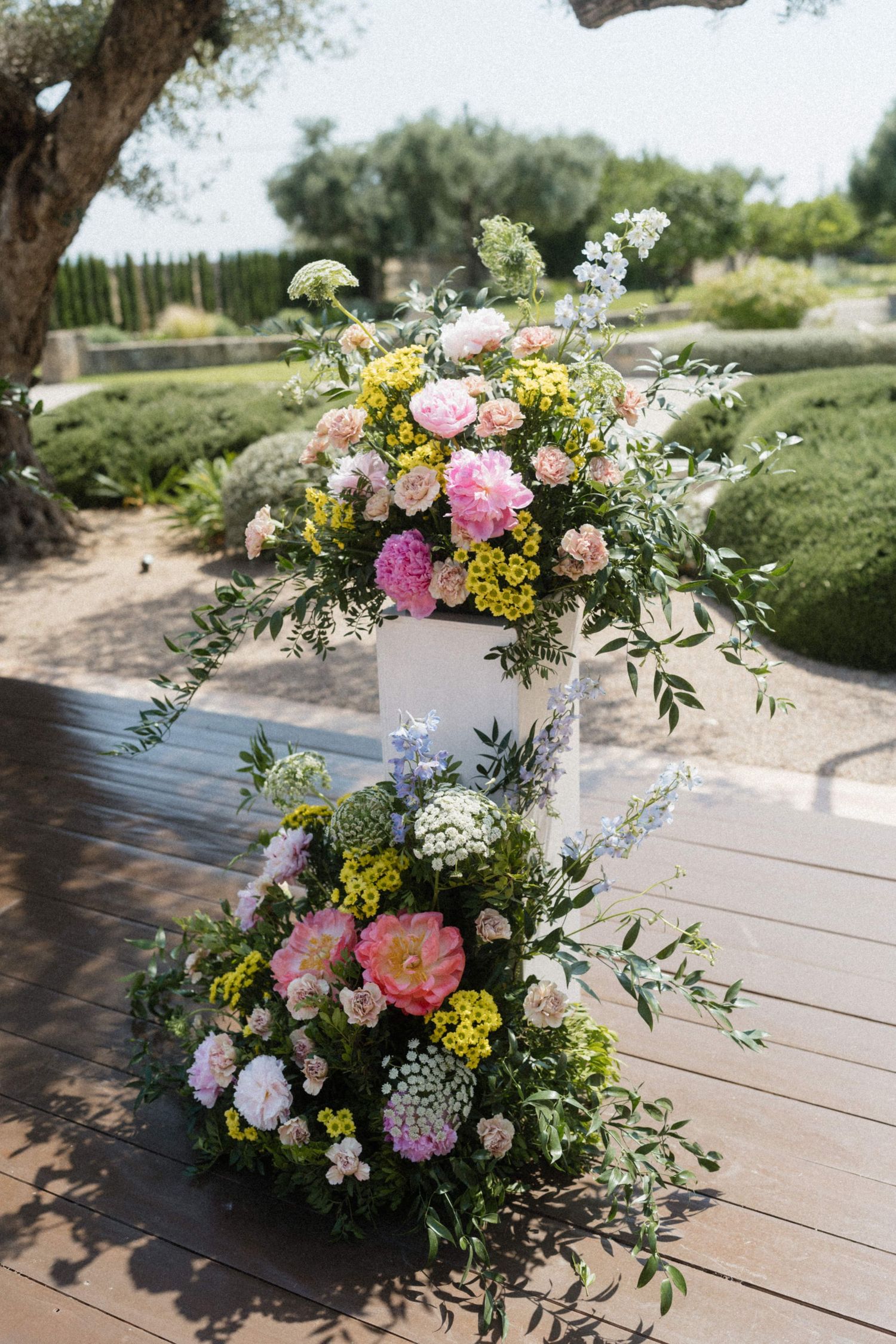 Elegant wedding ceremony floral arrangement with pink peonies, yellow flowers, and greenery on a wooden deck.