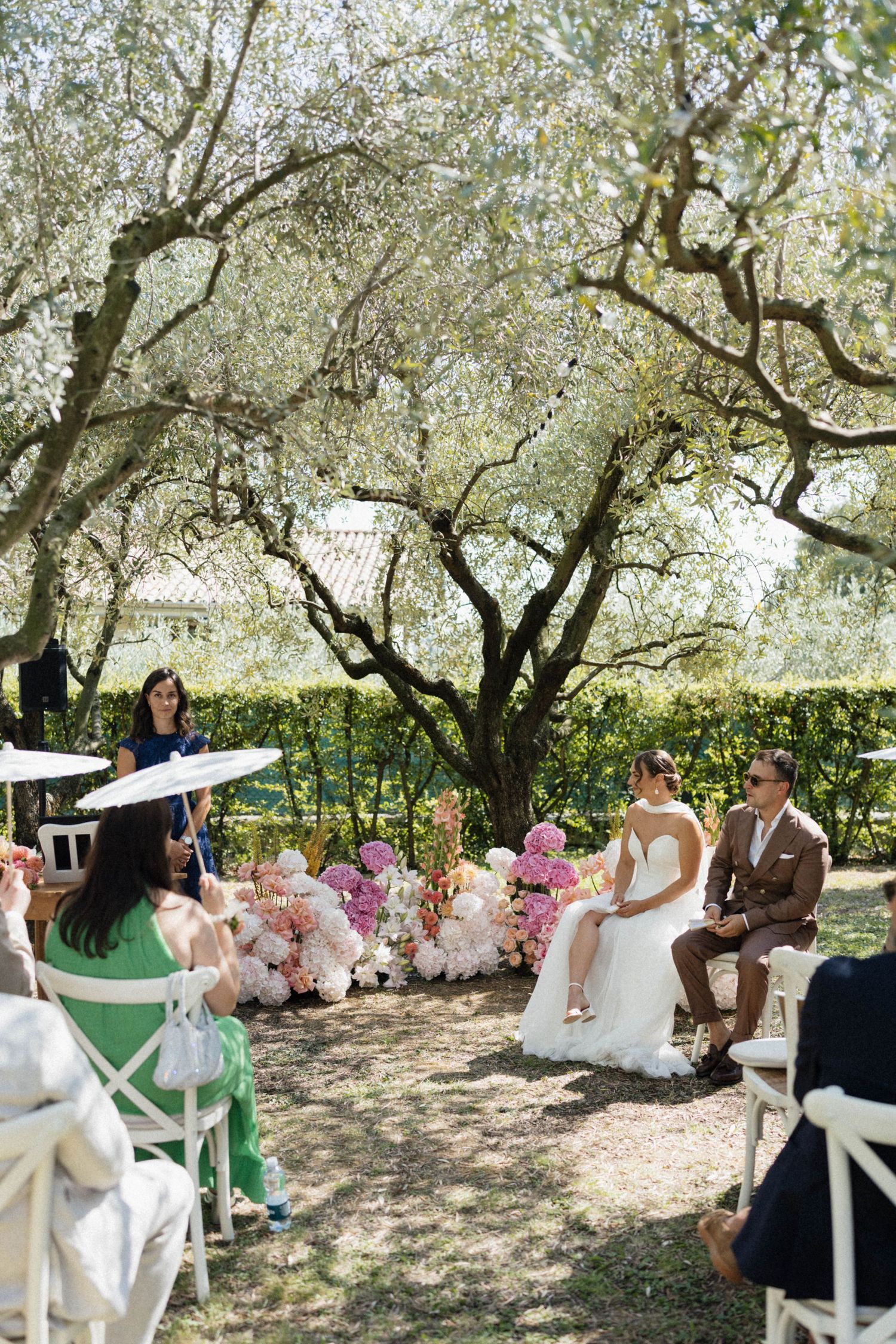 Outdoor wedding ceremony under olive trees with bride and groom seated, surrounded by pink floral arrangements and guests with parasols.