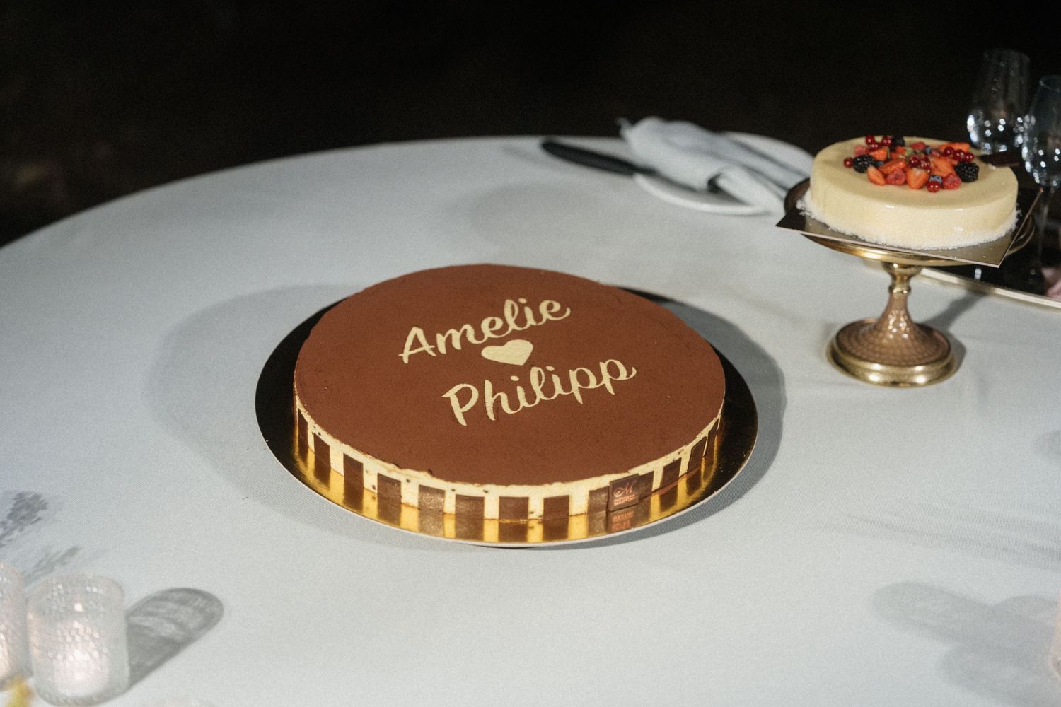Chocolate cake with gold trim inscribed with Amelie and Philipp on a white table at an elegant event.