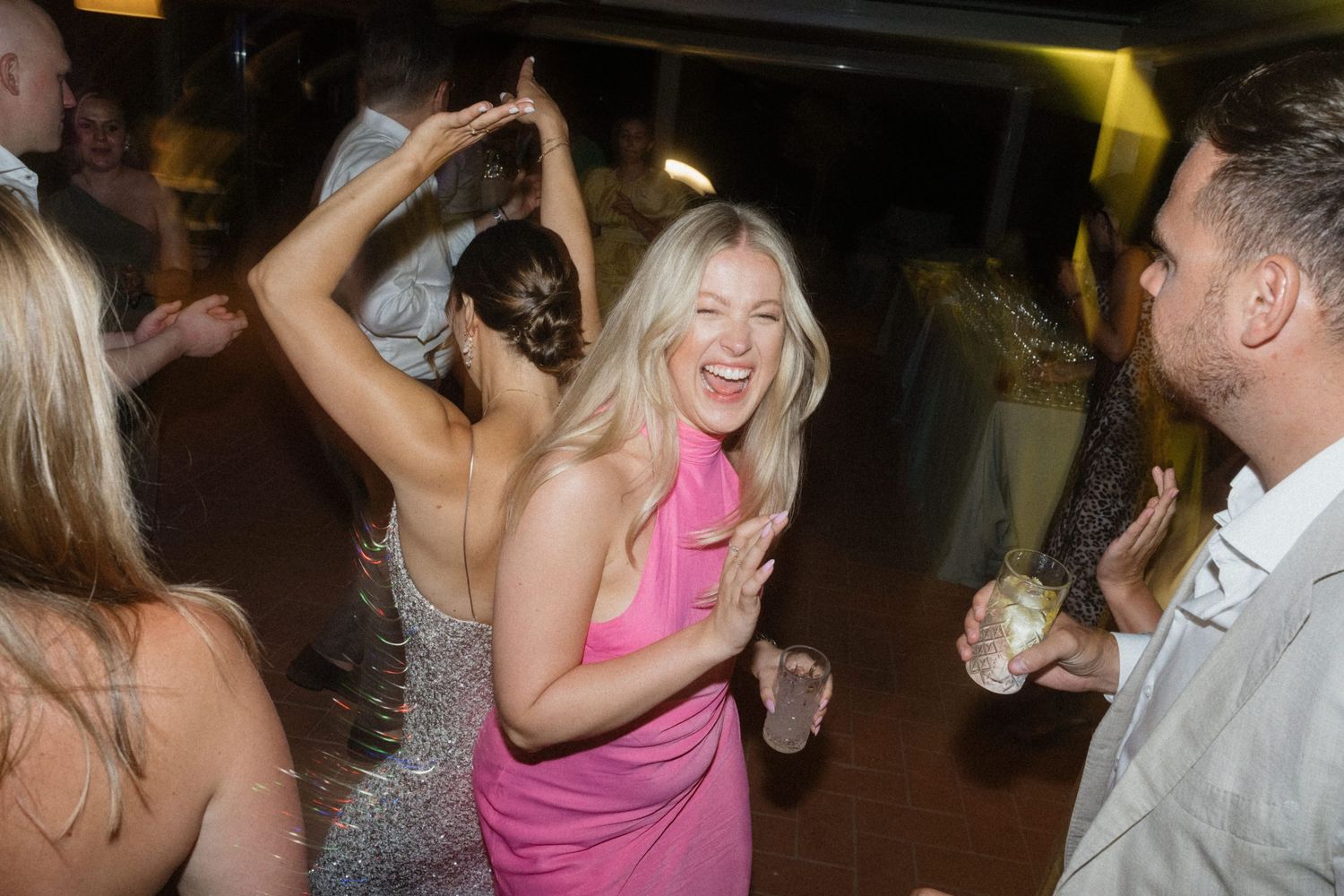 Guests dance joyfully at a wedding reception, woman in pink dress smiling.