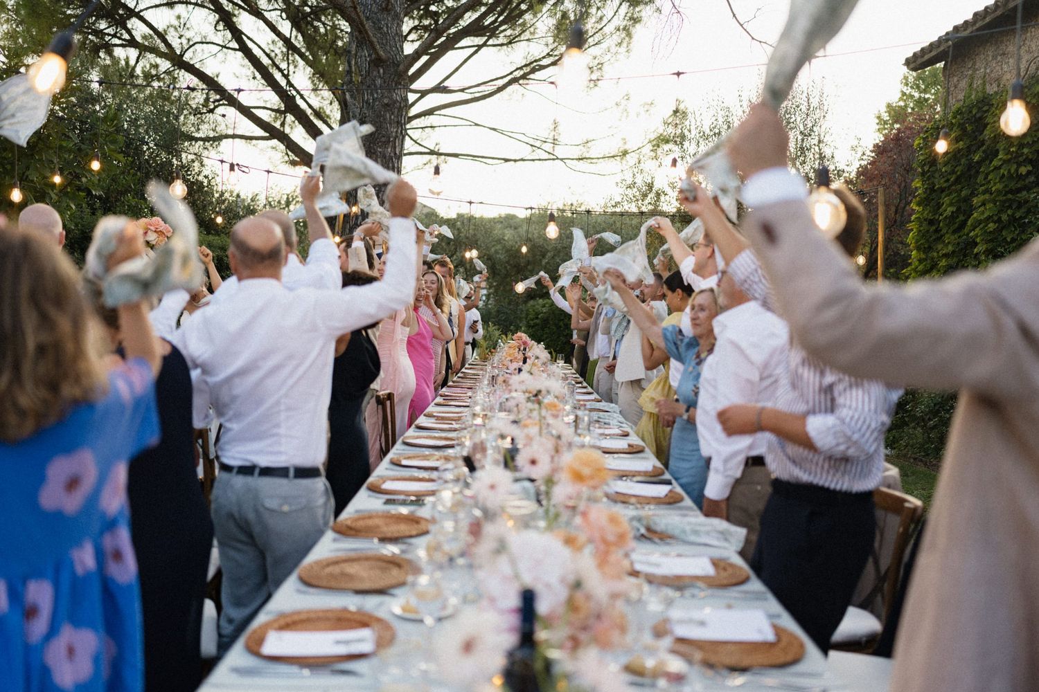 Guests celebrate at an elegant outdoor wedding reception with long decorated table, string lights, and raised napkins.