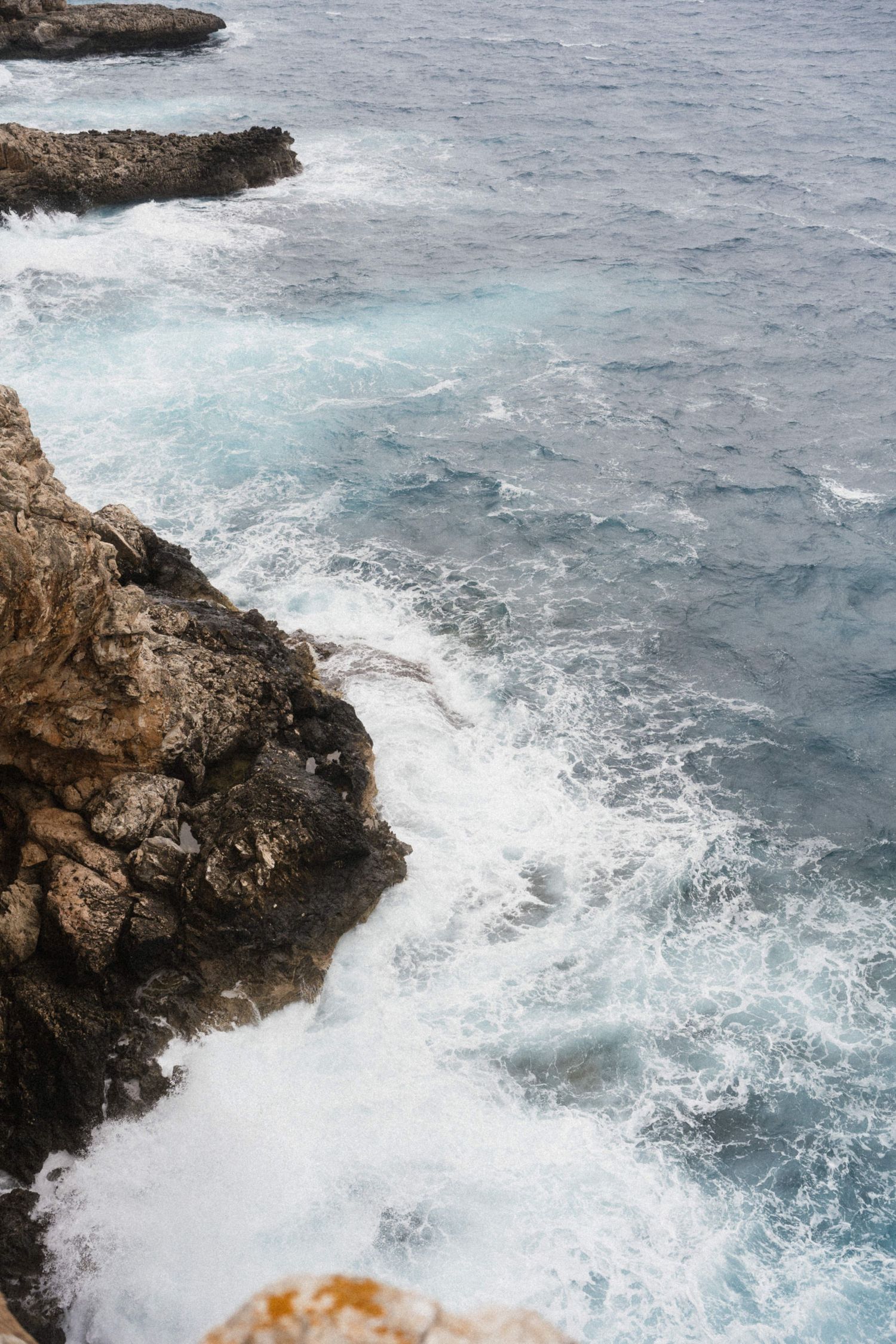 Rocky coastline with turquoise ocean waves crashing against rugged cliffs, creating white sea foam on a cloudy day.