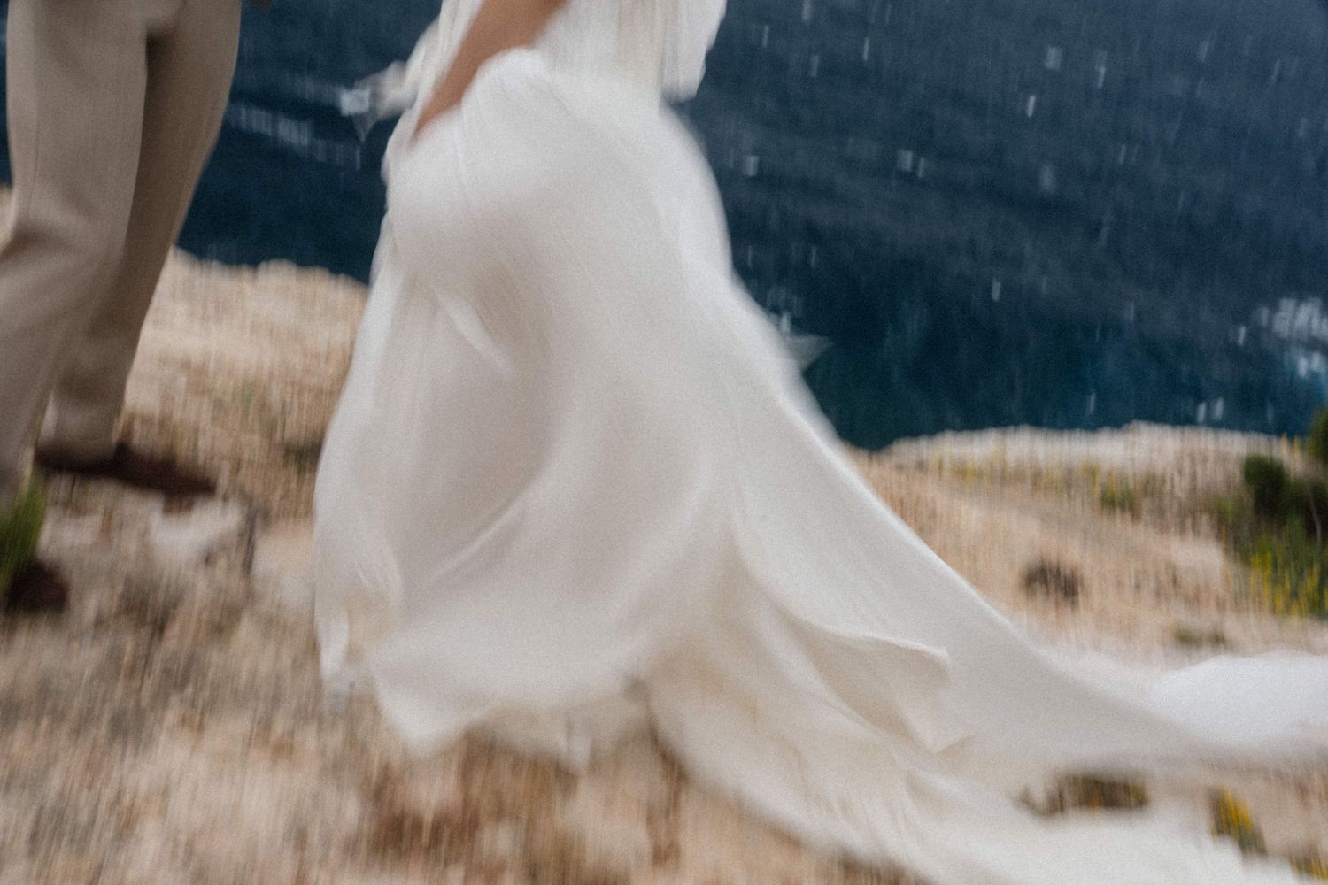 Bride in flowing white wedding gown with long train walks outdoors on a cliffside in the rain.