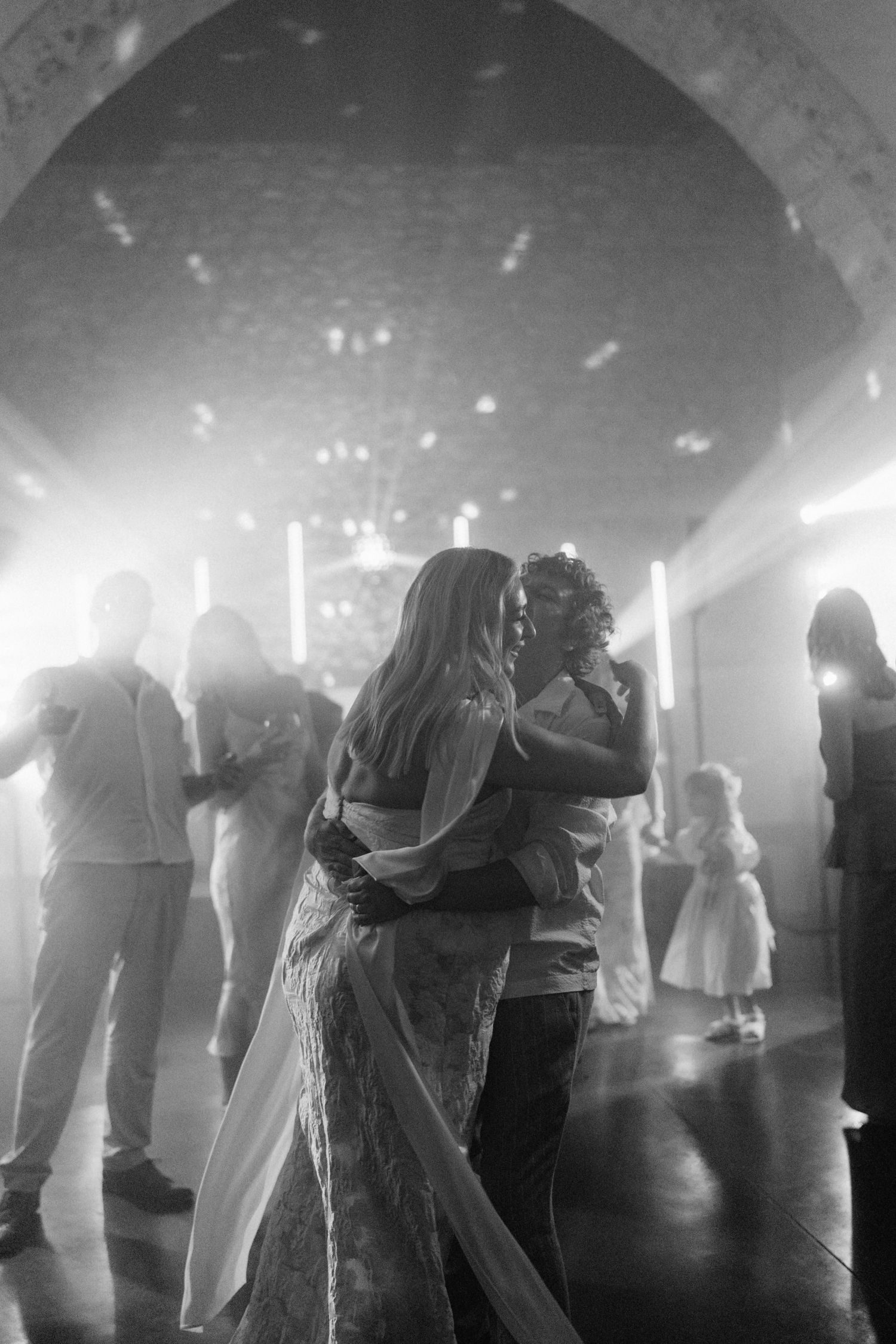 Black and white photo of couple sharing first dance at wedding reception under arched ceiling with soft glowing lights.