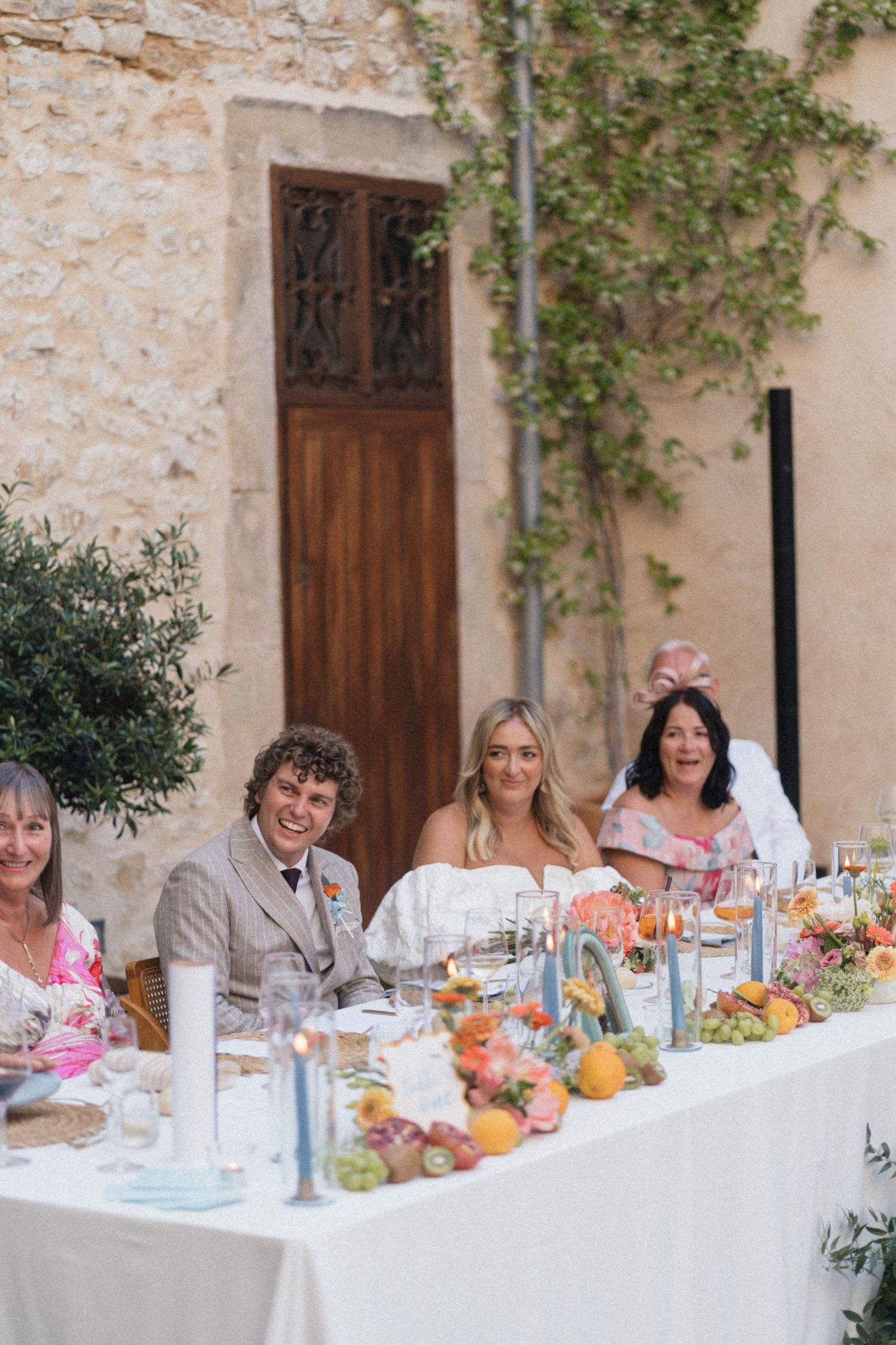 Smiling bride and groom sit at a beautifully decorated wedding reception table adorned with colorful fruit and flowers.