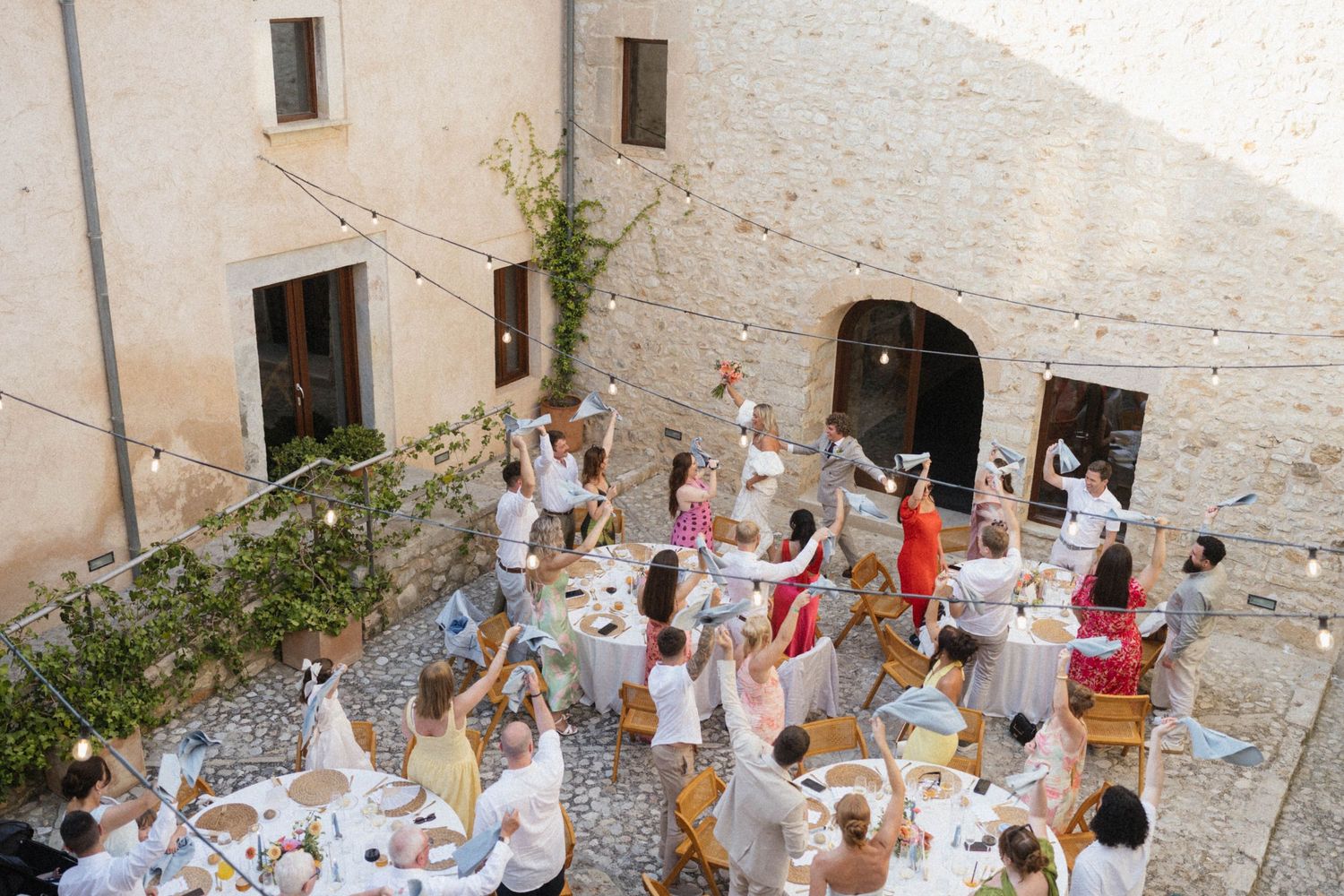 Guests raising glasses in a toast at an outdoor courtyard wedding reception with string lights in Italy.