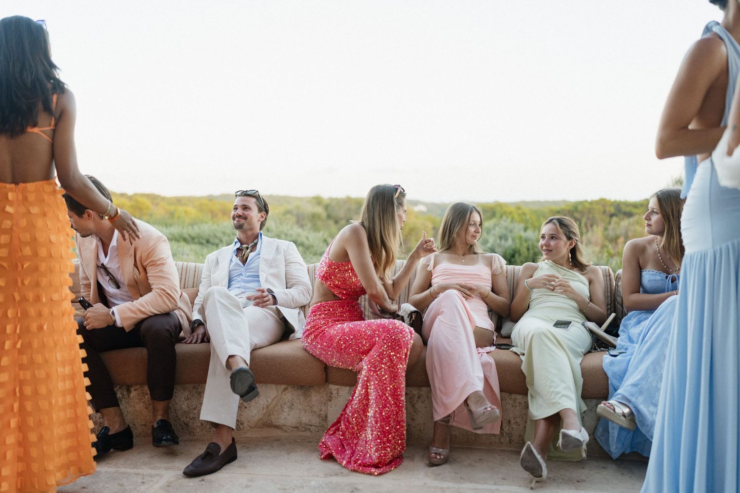 Wedding guests in colorful dresses and suits chat on a stone bench at a sunset outdoor celebration.