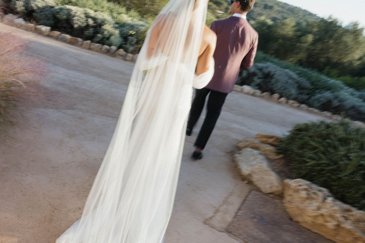 Bride in a long flowing veil and gown walks hand in hand with the groom along a stone path lined with shrubs.
