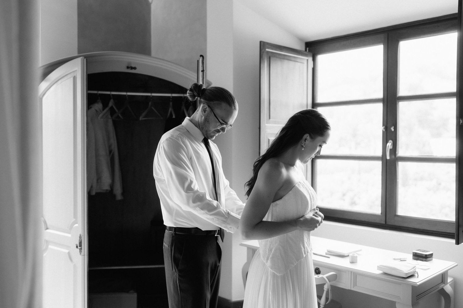 Black and white photo of a man helping a bride button her wedding dress beside a bright window in a cozy room.