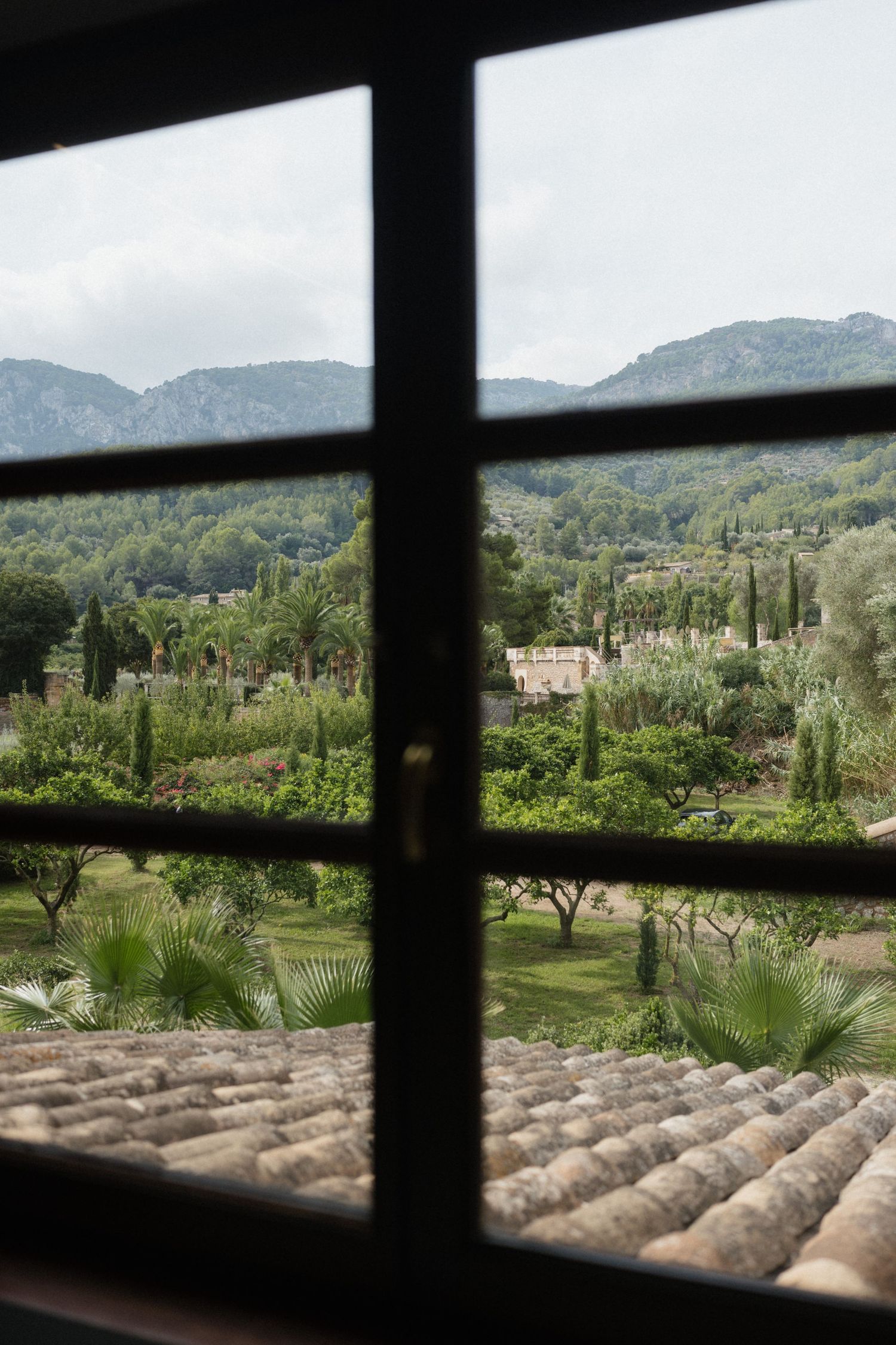 View through a dark-framed window of terracotta roof tiles, lush Mediterranean gardens, palm trees and misty mountains beyond.