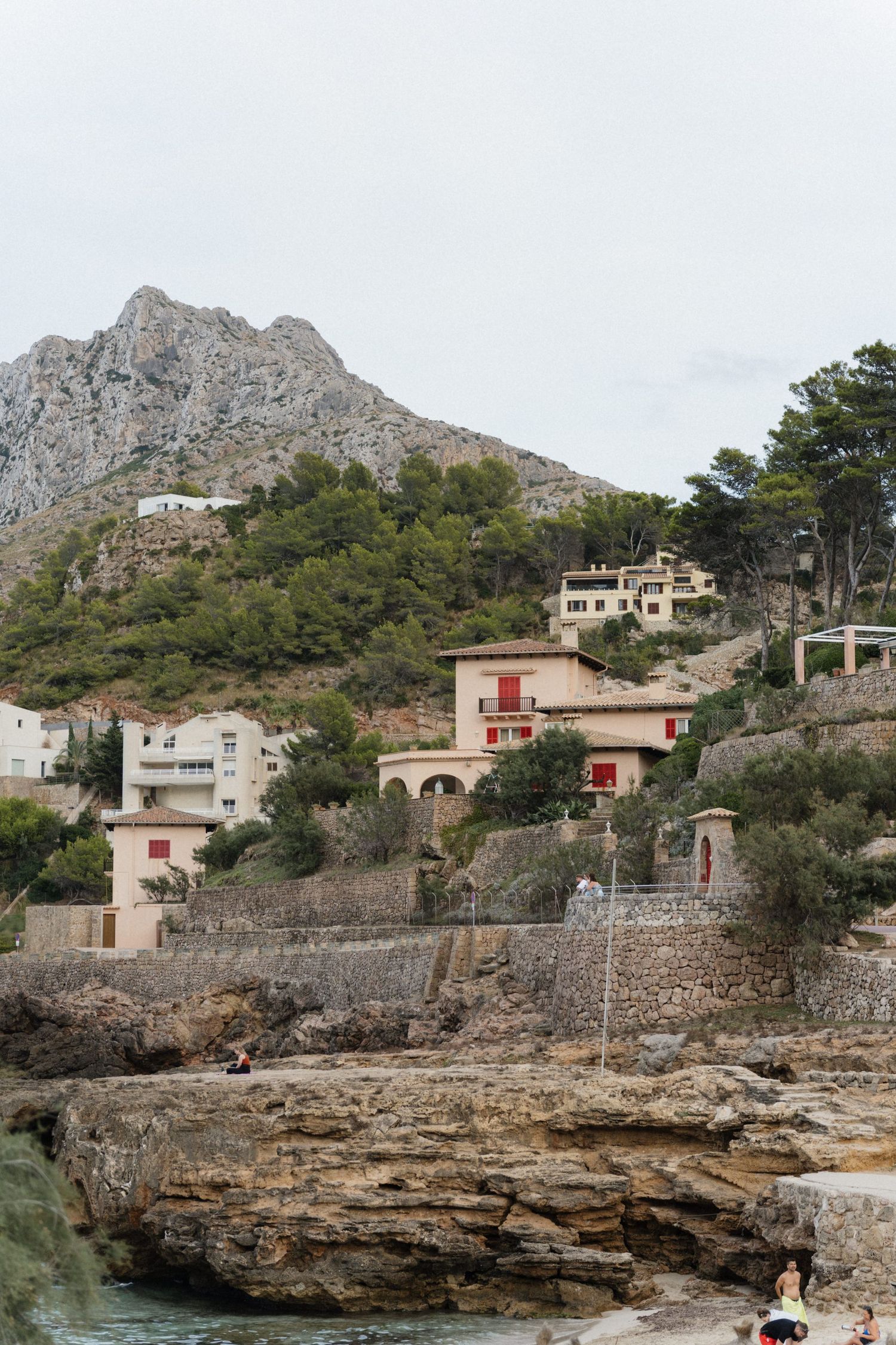 Mediterranean villas with red shutters perch on a rocky coastal hillside beneath a rugged mountain, with pine trees and a small cove below.