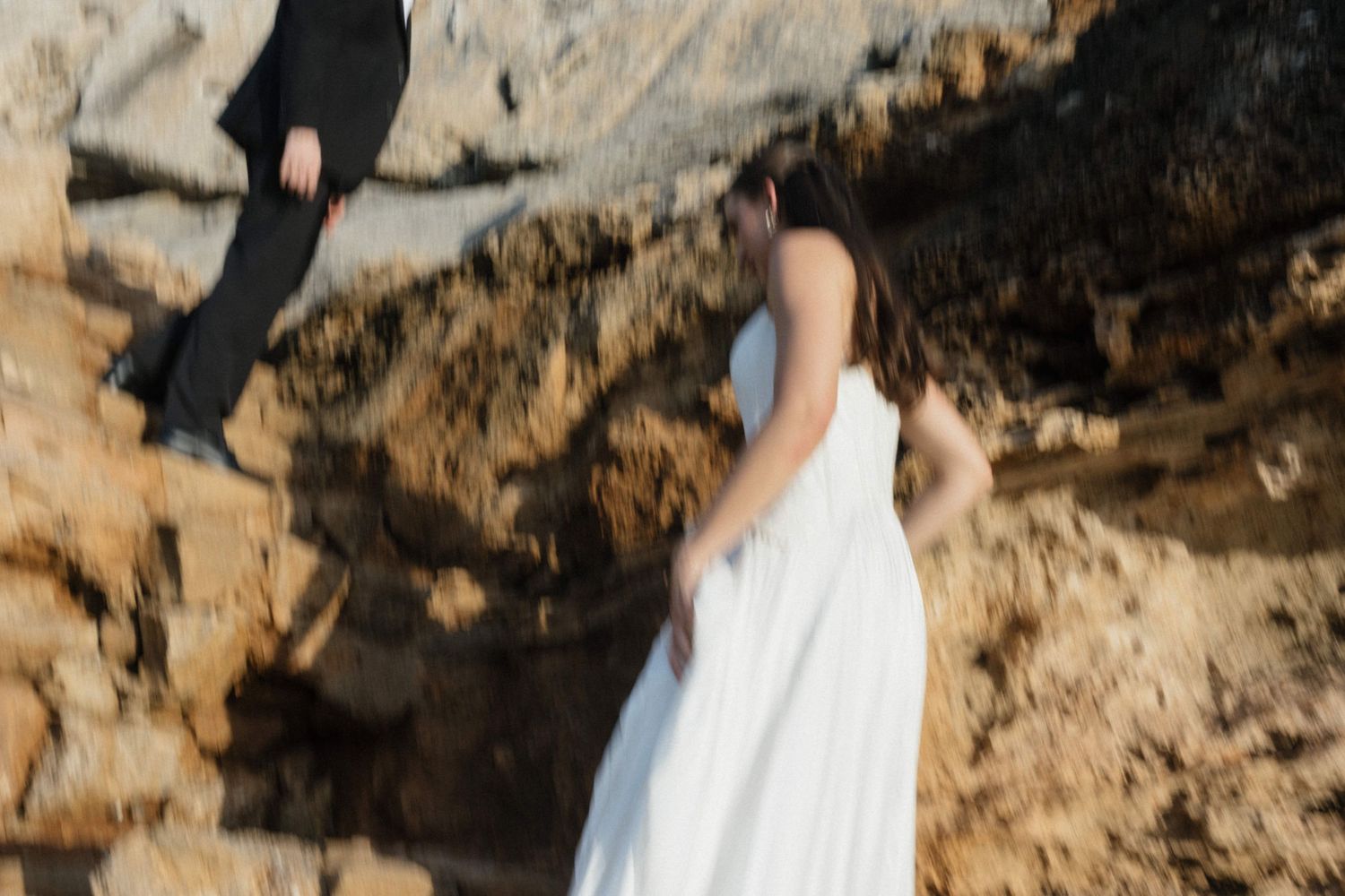 Bride in a white gown walks along orange coastal rocks while the groom in a black suit stands behind her.