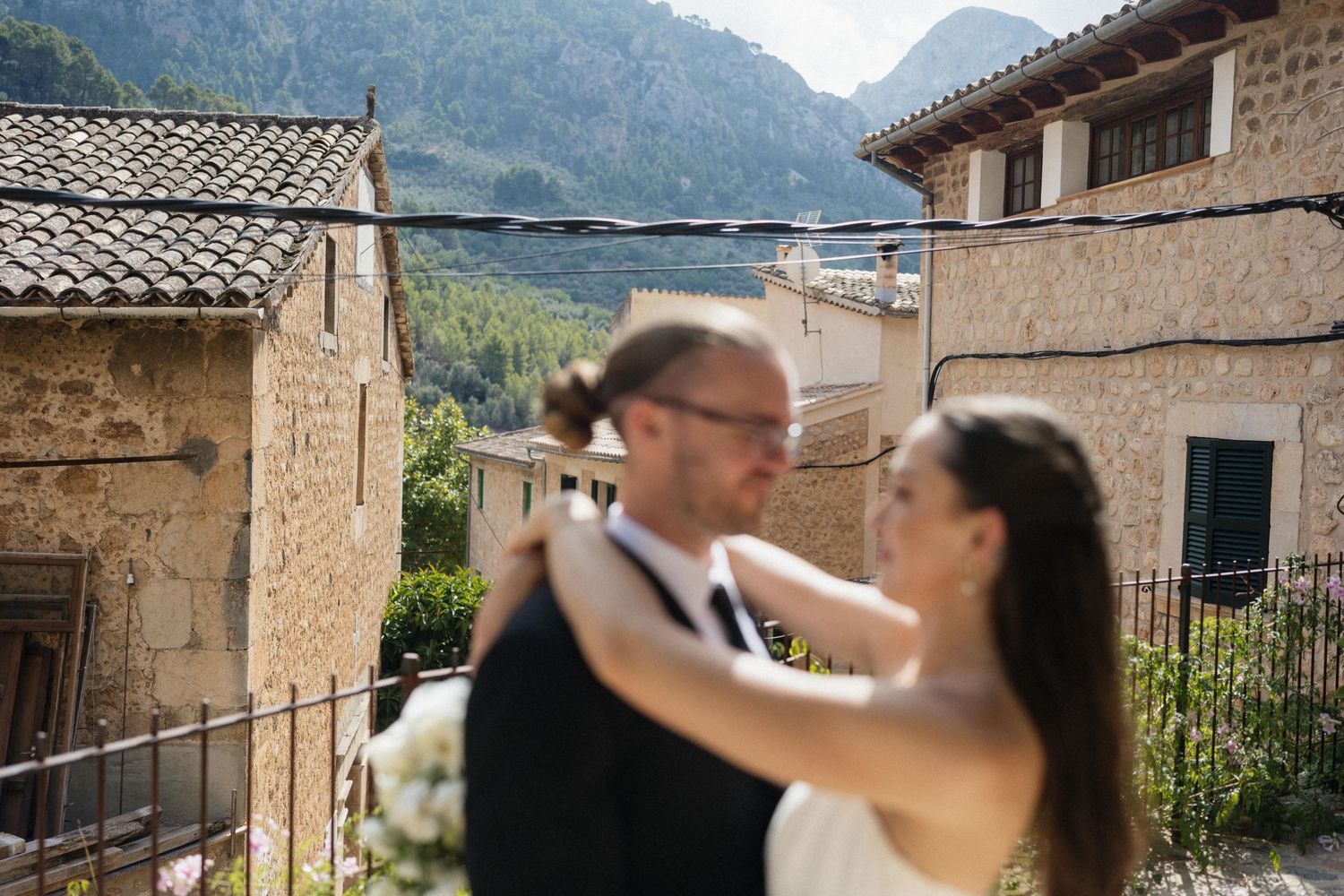 Bride and groom embrace in a romantic wedding portrait with a rustic stone village and forested mountains in the background.