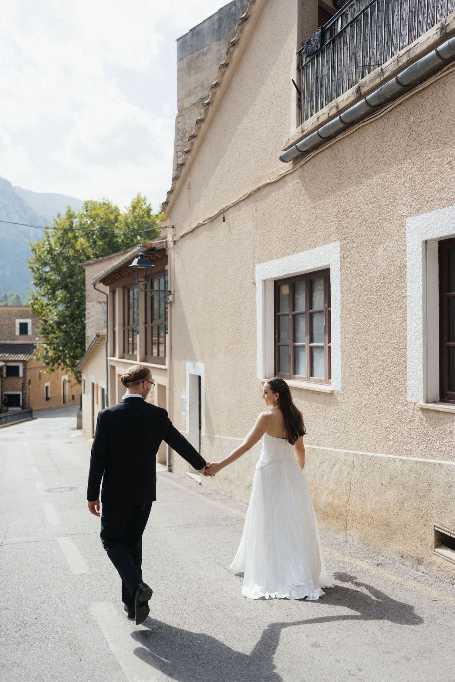 Bride in a flowing white gown and groom in a dark suit walk hand in hand down a sunlit village street.