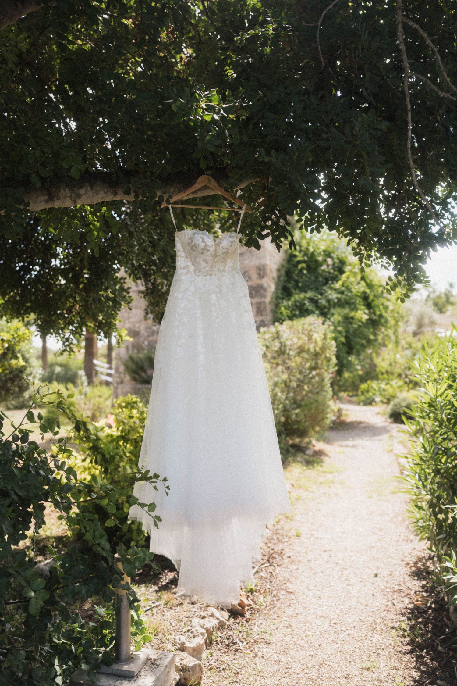 Elegant white lace wedding gown hanging on wooden hanger from tree in garden pathway.