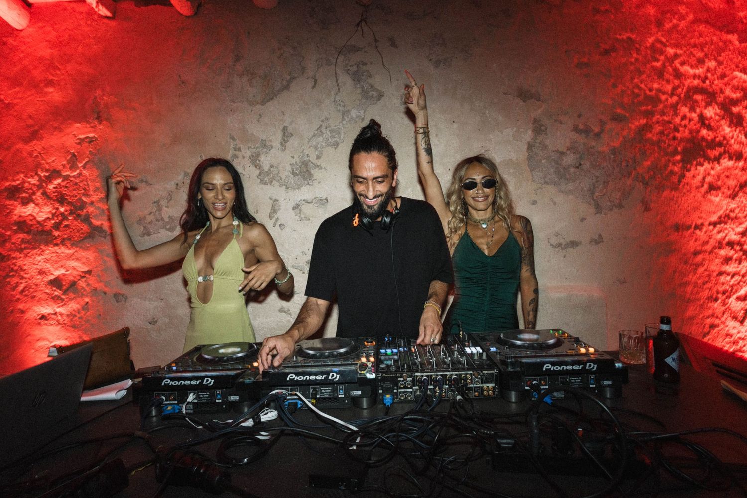DJ performing at wedding reception flanked by two women dancing under red ambient lighting.