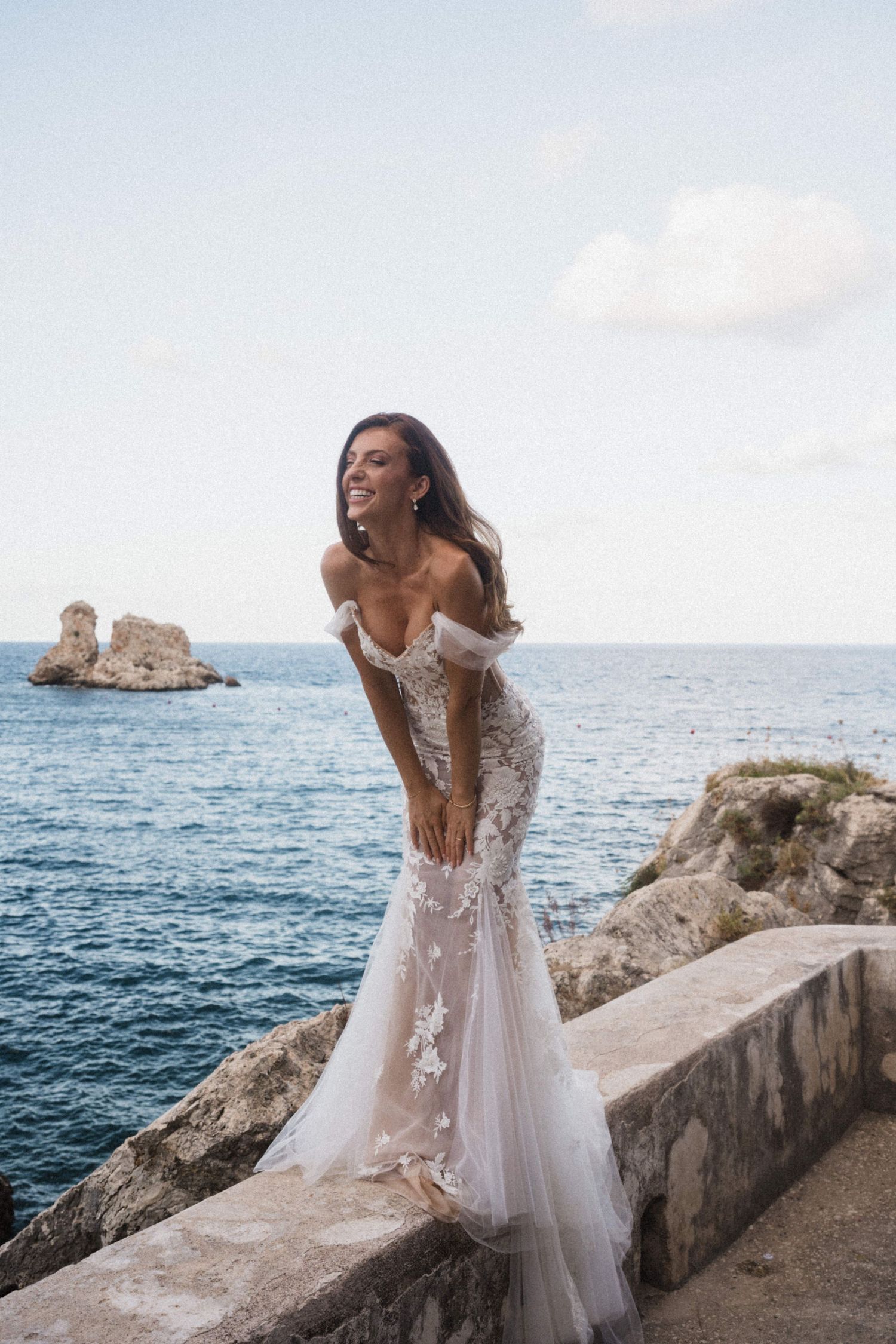 Radiant bride in off-shoulder lace mermaid wedding dress smiling on rocky coastal terrace overlooking blue sea.