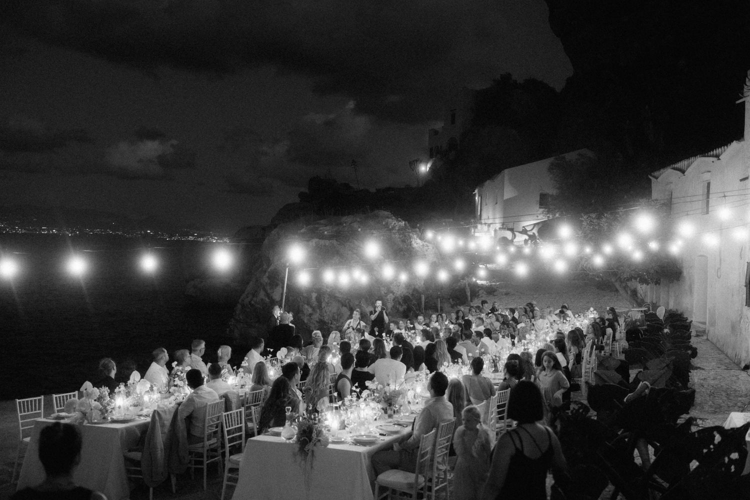 Black and white aerial view of a stunning outdoor wedding reception with string lights overlooking a city at night.