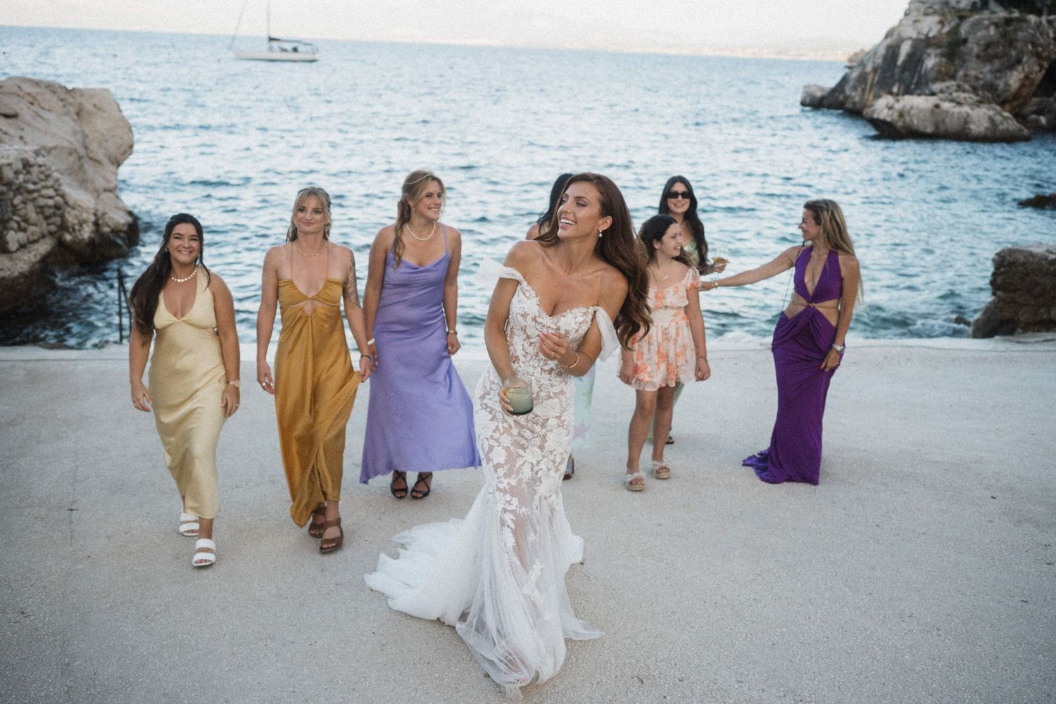 Bride in lace gown walks with colorful bridesmaids by the sea at a coastal wedding.