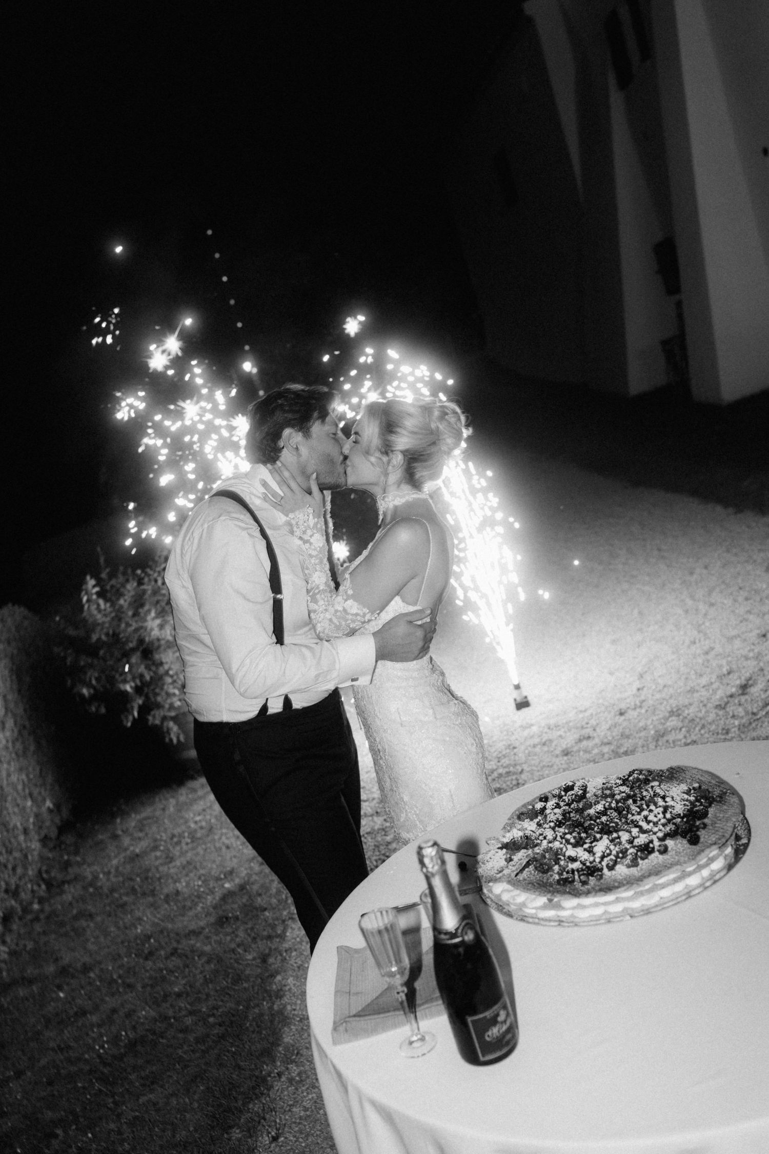 Black and white photo of newlyweds kissing beside wedding cake with sparklers glowing in background.
