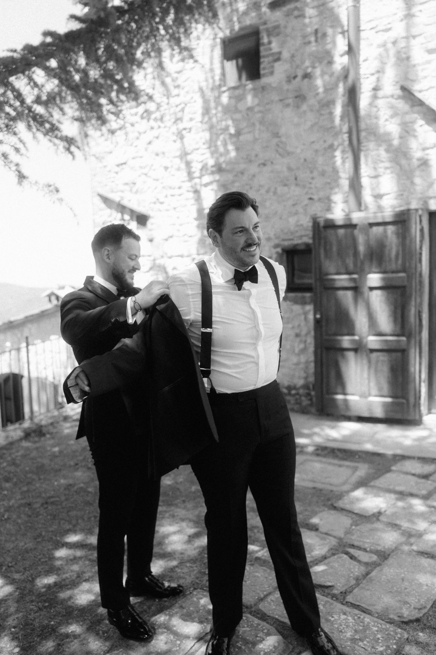 Black and white photo of groomsman helping groom put on tuxedo jacket outside stone building.