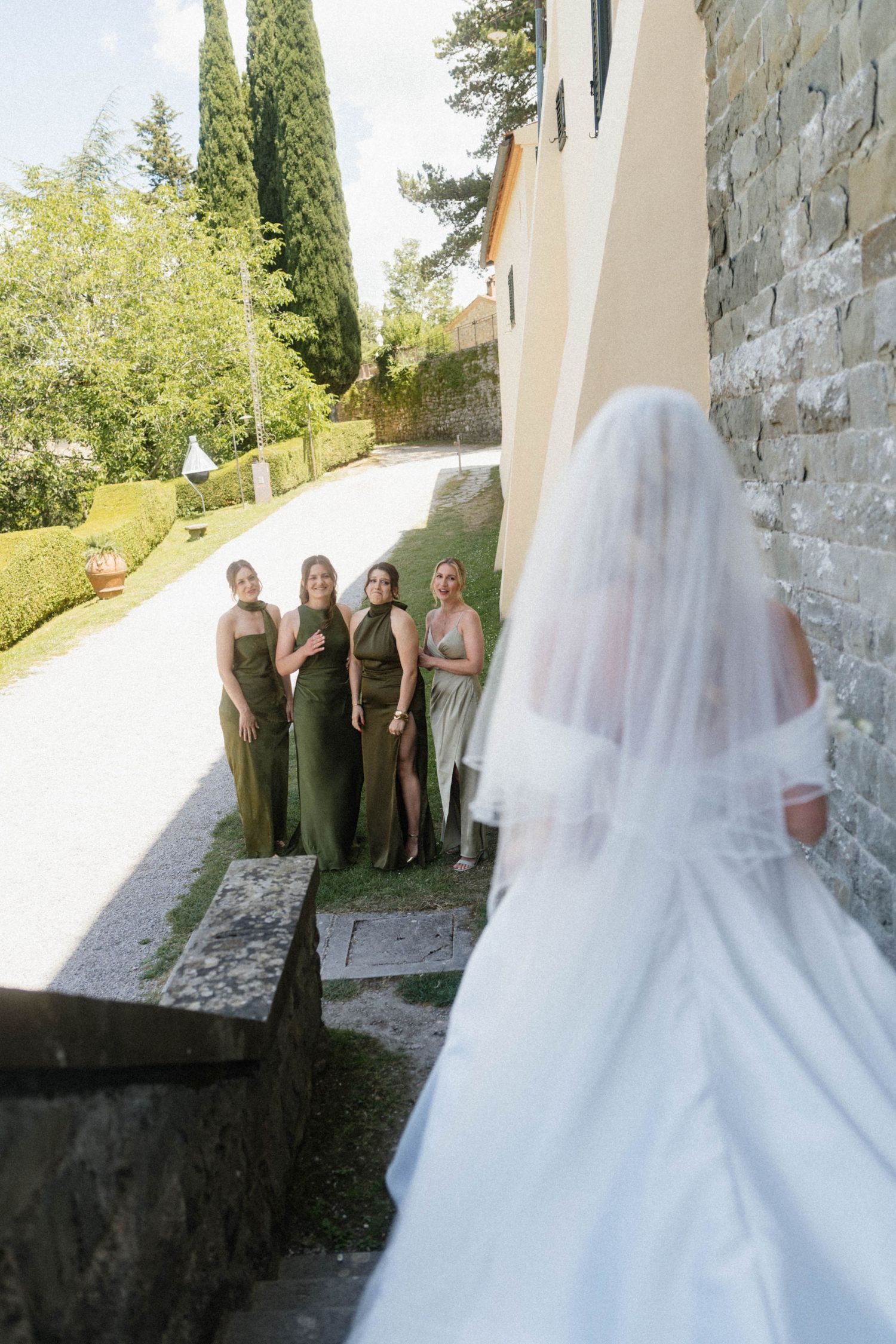 Bridesmaids in olive green dresses reacting with joy as bride in white veil approaches.