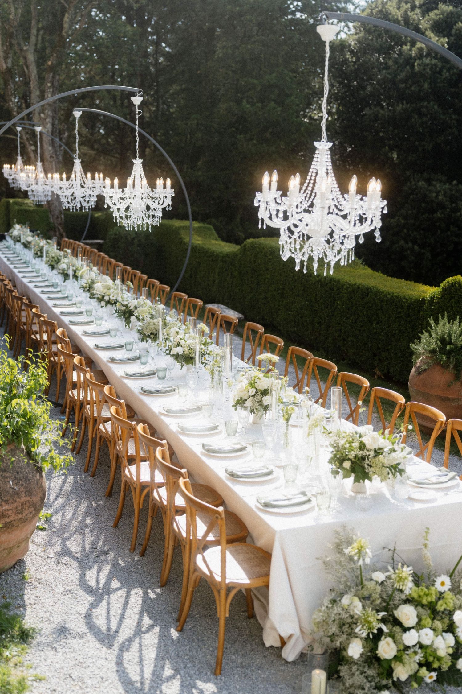 Stunning outdoor wedding reception with long banquet tables, crystal chandeliers, and white floral arrangements.