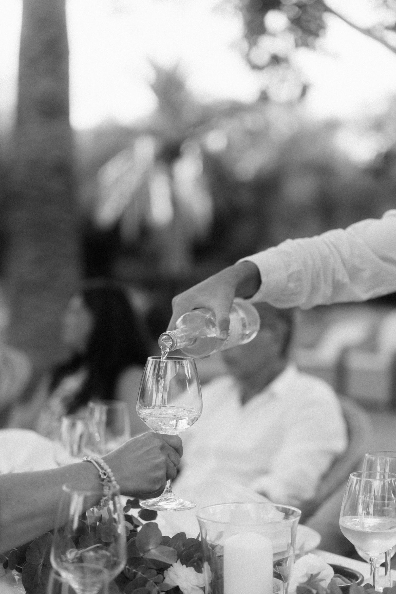 Black and white photo of waiter pouring white wine into guest glass at outdoor wedding.