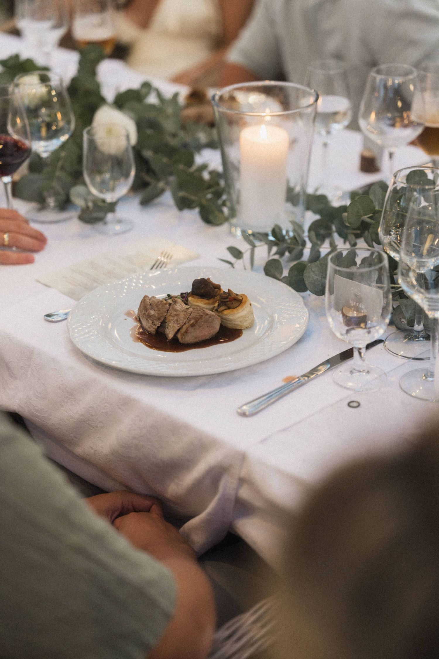 Elegant wedding dinner plate with meat dish beside candles and eucalyptus greenery centerpiece.