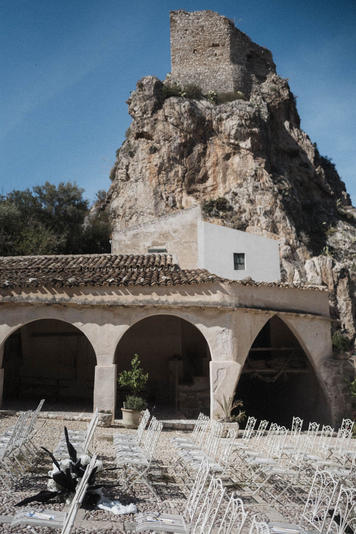 Ancient stone chapel with arched portico and white chairs set for outdoor wedding ceremony beneath dramatic rocky cliff.