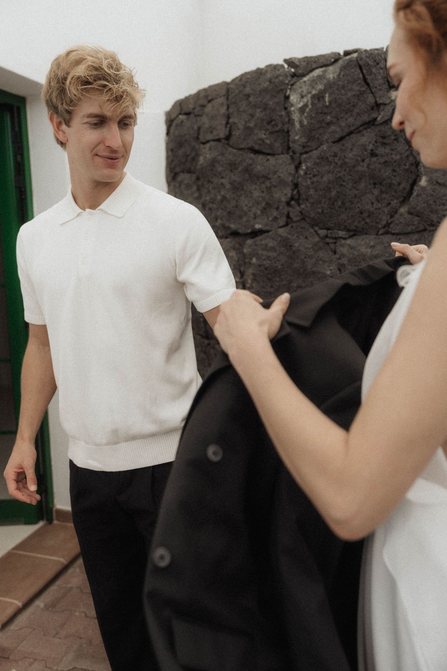 Groom in white polo smiles at bride holding black jacket near volcanic rock wall.