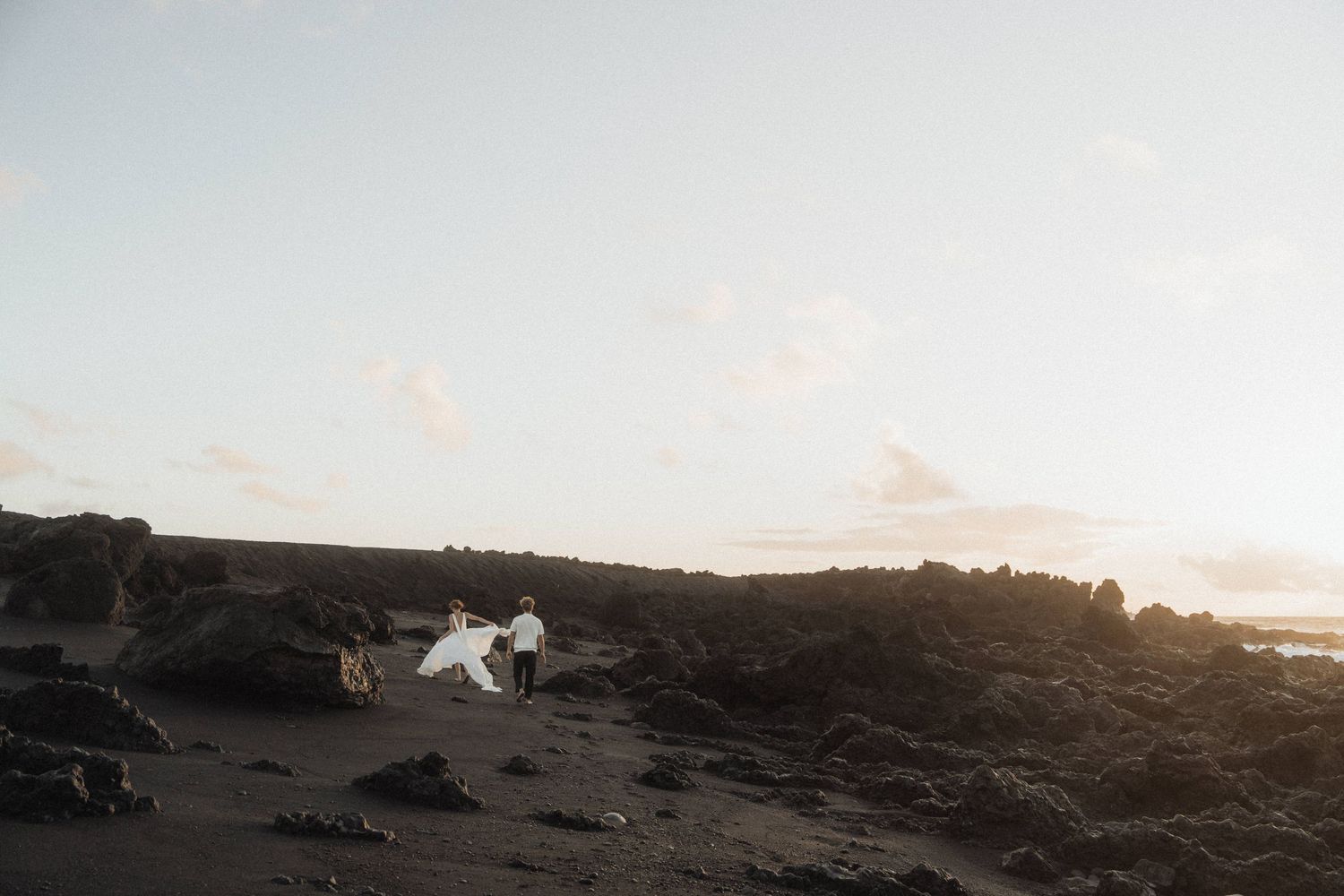 Bride and groom walking across dramatic black volcanic landscape under a soft pastel sunset sky.