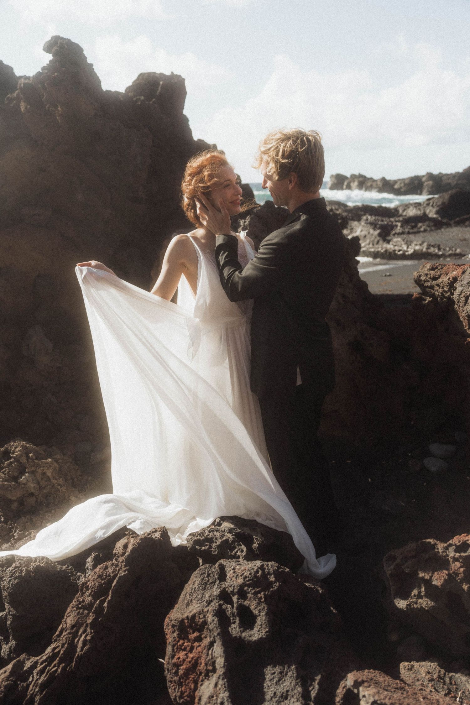 Bride and groom embrace on rocky coastal cliffs, her white flowing dress billowing in the ocean breeze.