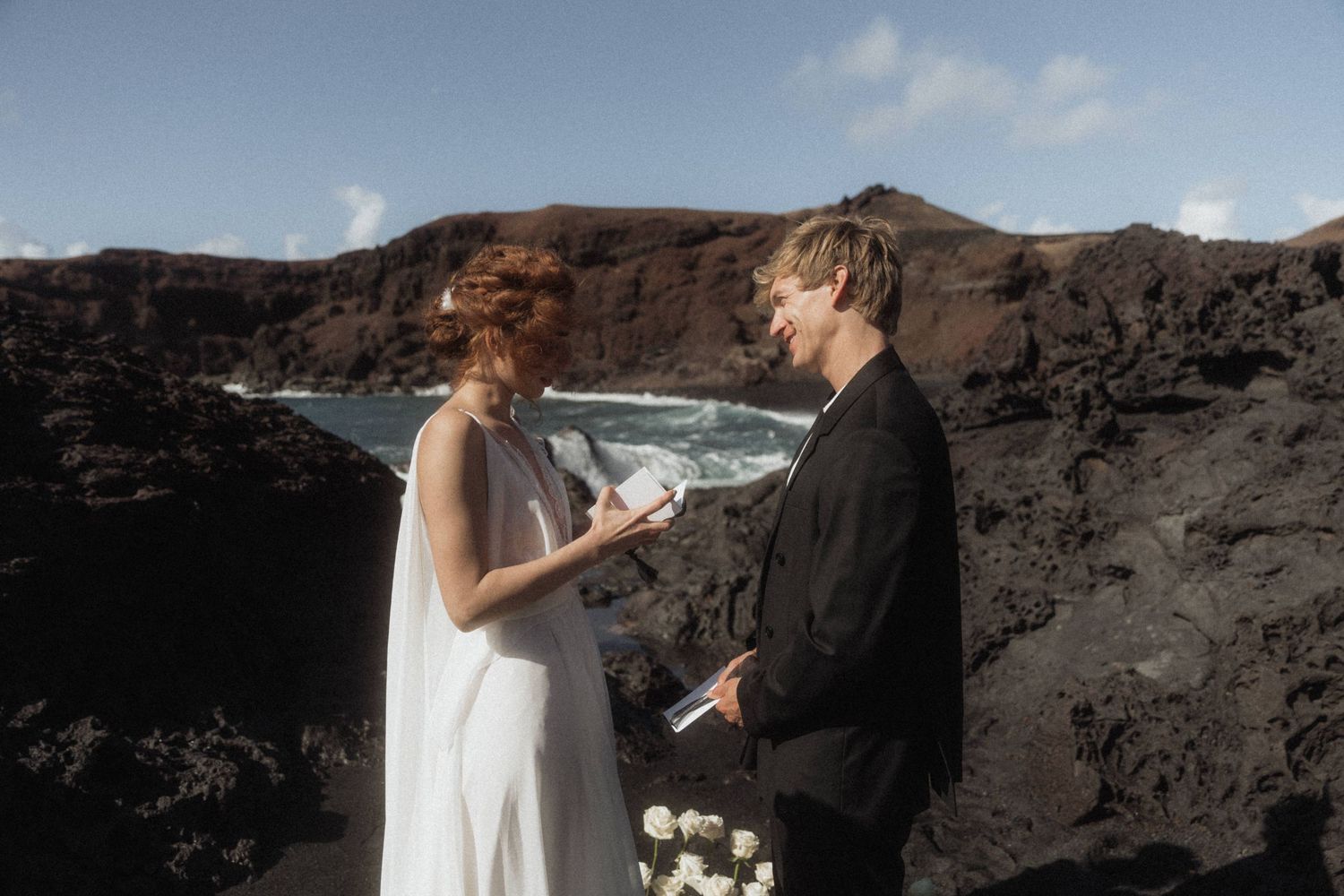 Couple exchanging vows at a dramatic volcanic cliffside wedding, bride in white gown holding vows, groom in black suit.