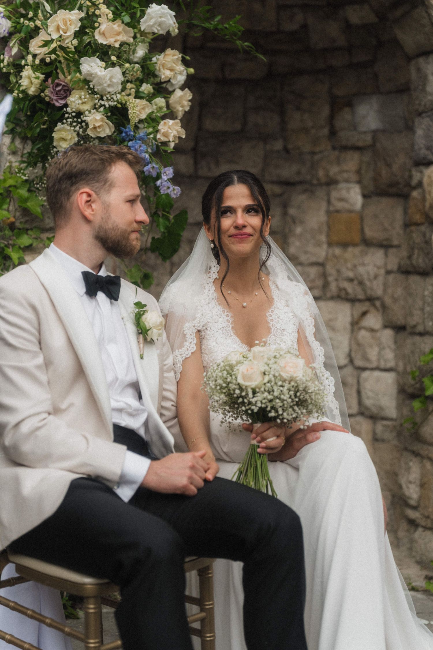 Bride in lace veil holding white bouquet seated beside groom at wedding ceremony.