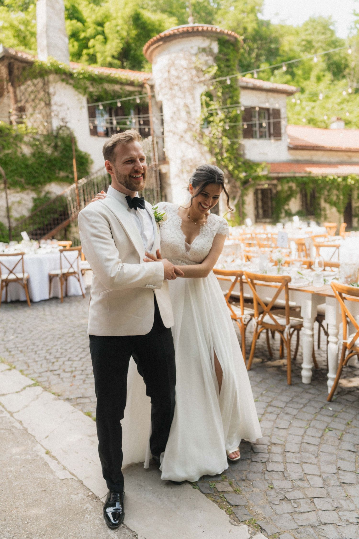Bride and groom share a joyful moment in a cobblestone courtyard with elegant outdoor reception tables and lush greenery.