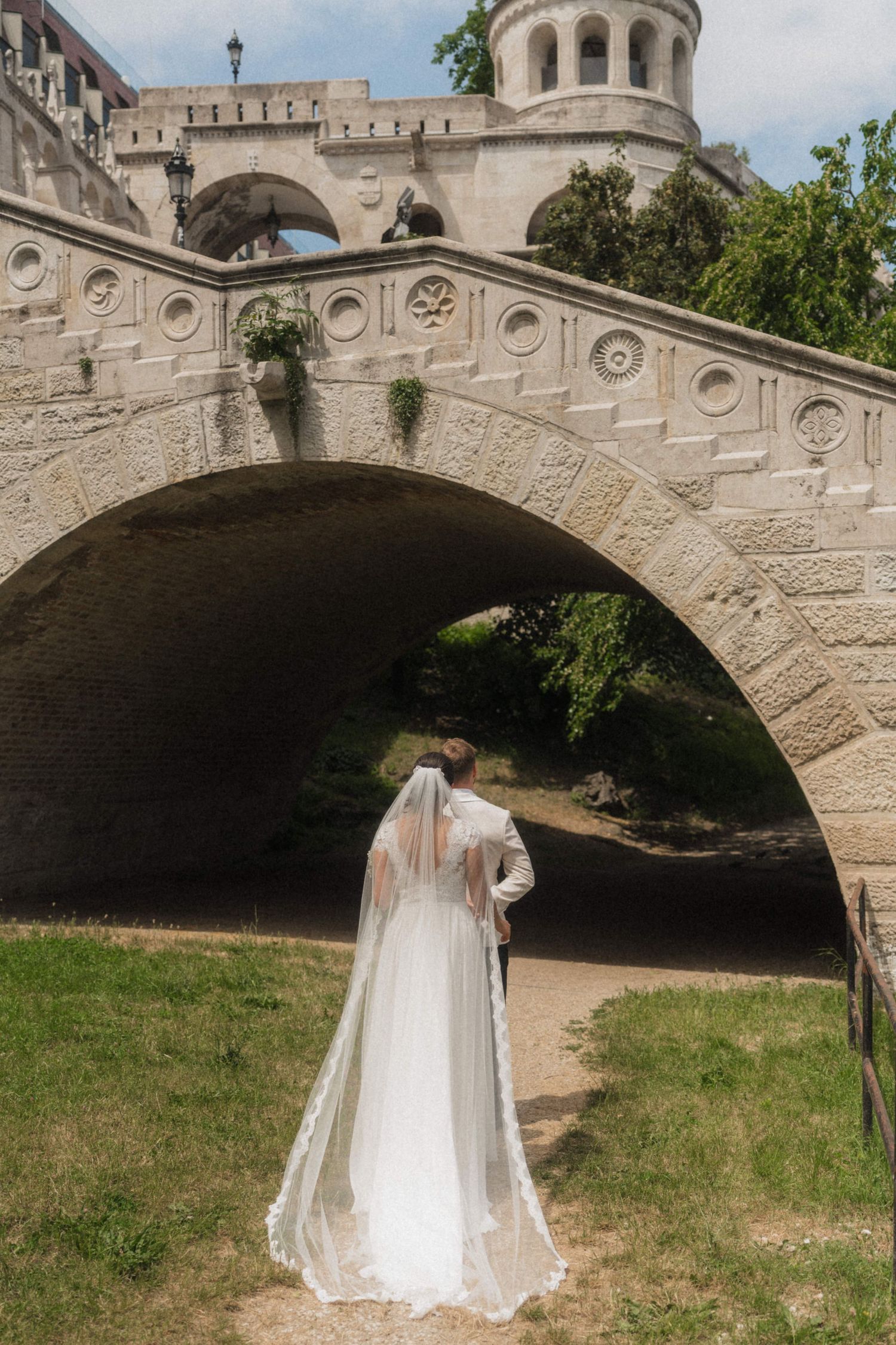 Bride and groom walking hand in hand toward a stone arch bridge on their wedding day.