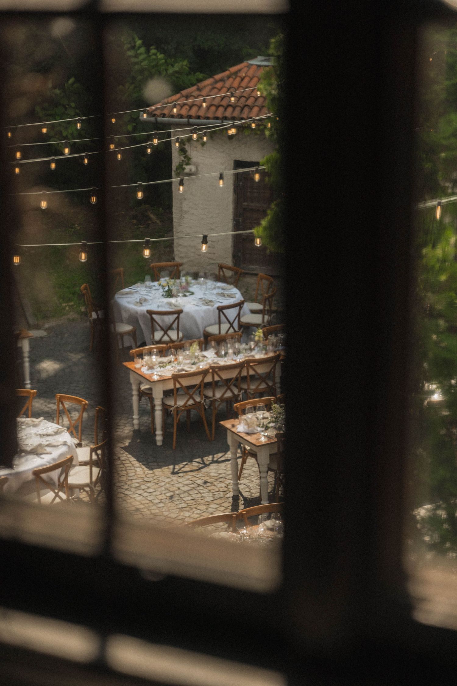 Romantic outdoor wedding reception viewed through window with string lights and rustic wooden chairs.
