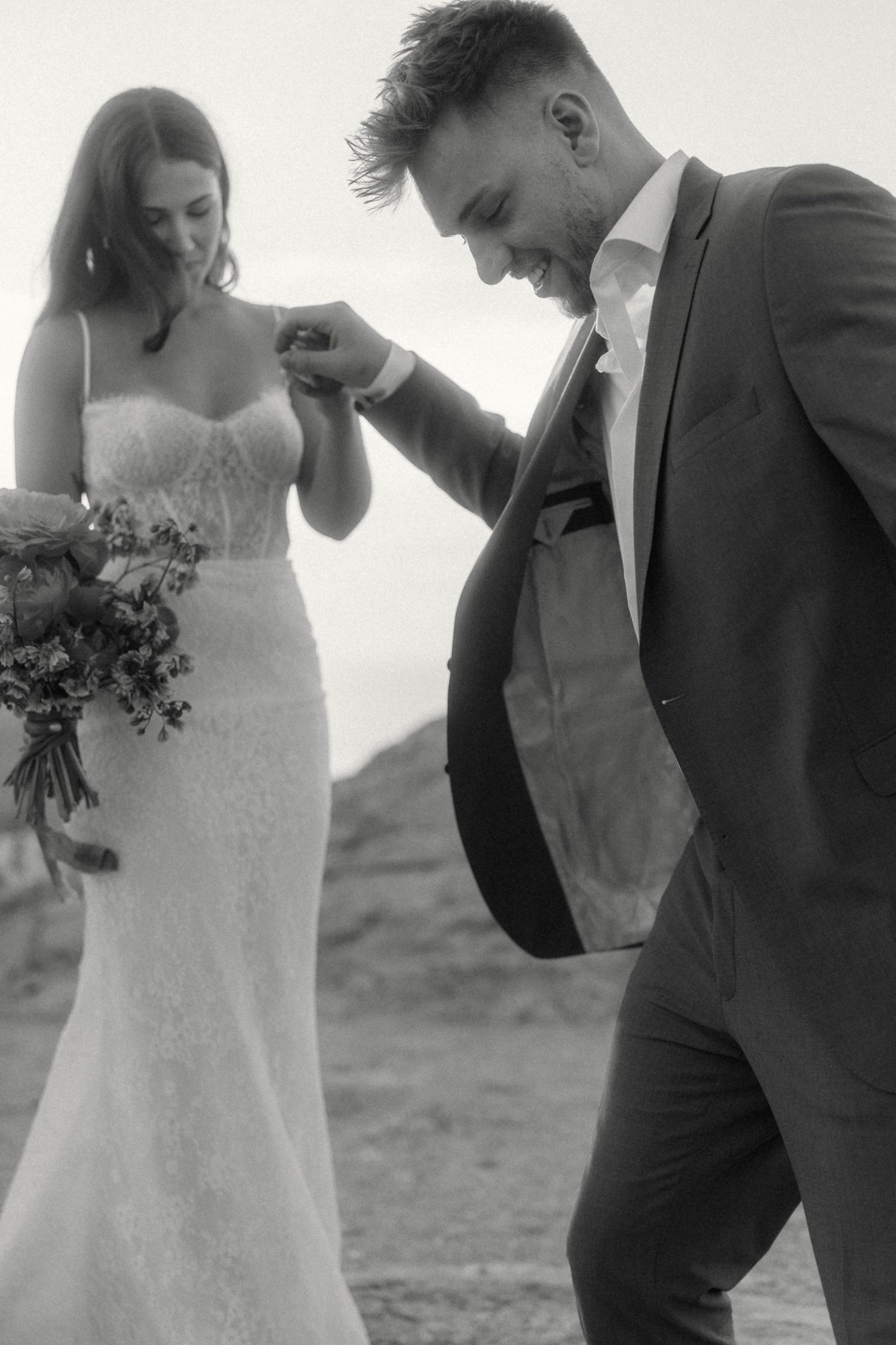 Black and white photo of a smiling groom leading his bride by the hand, bride holding a floral bouquet.