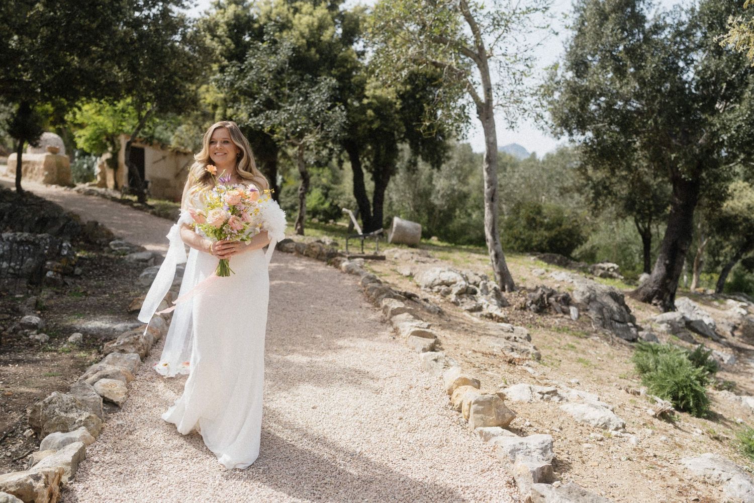 Bride in white gown holding floral bouquet walks along stone path surrounded by olive trees at outdoor wedding.