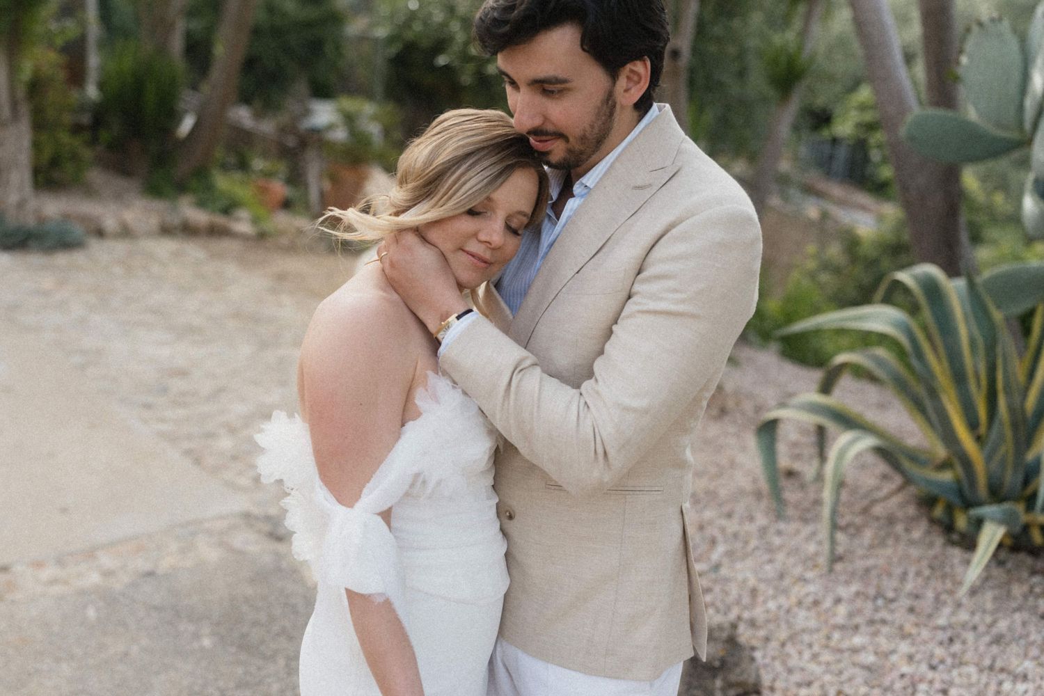 Groom in beige linen suit tenderly embracing bride in white feather wedding dress outdoors.