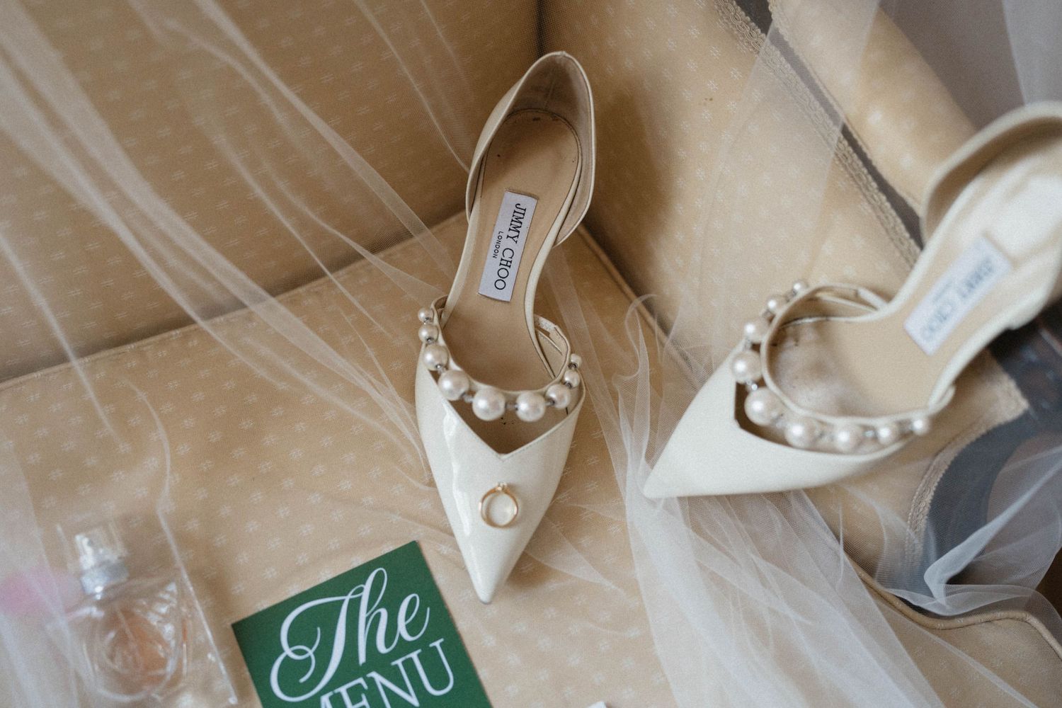 Elegant ivory Jimmy Choo bridal heels with pearl embellishments displayed on a chair with a wedding veil and menu card.